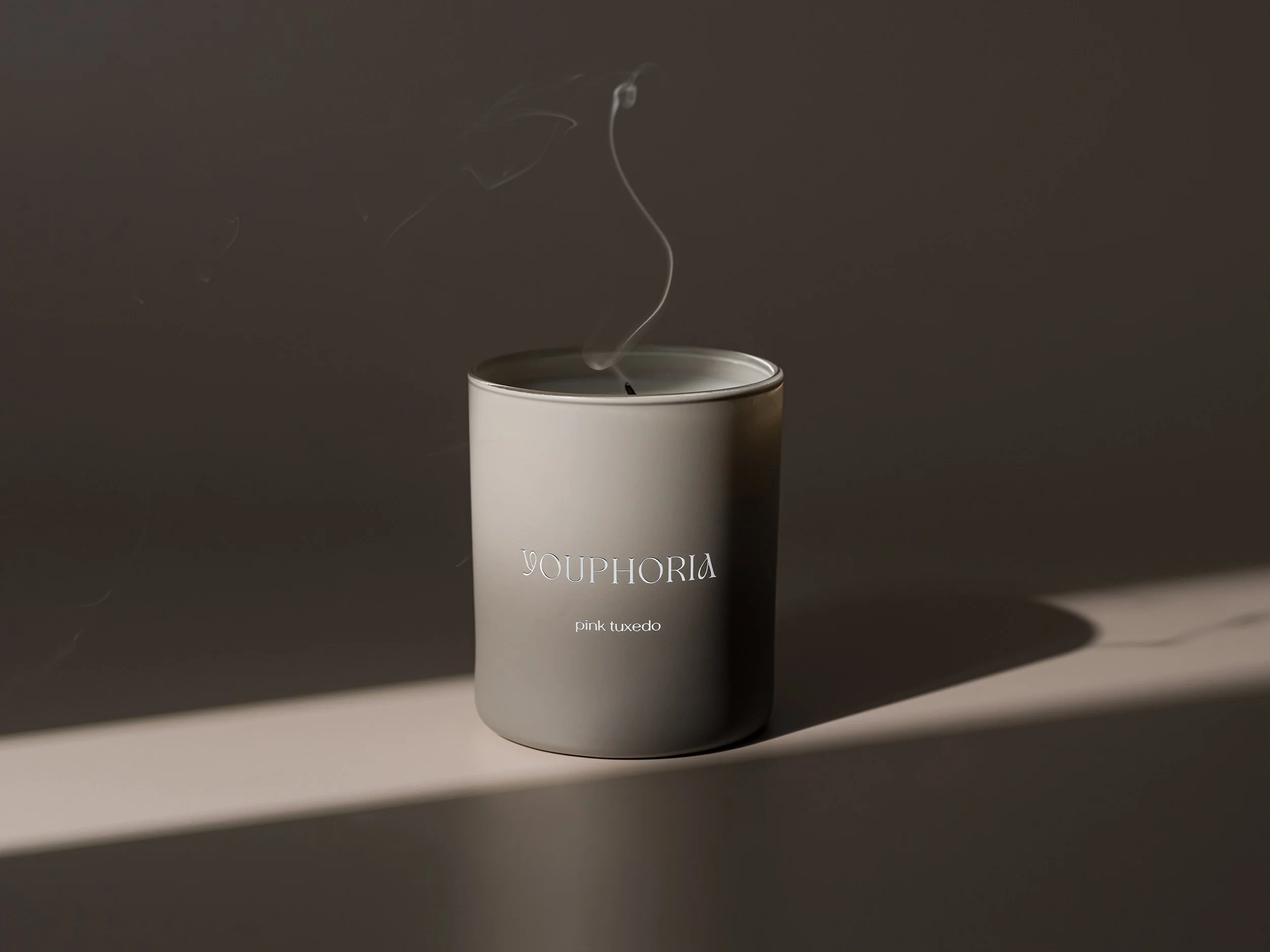 A gray candle with white text that reads "YOU PHORIA" and "pink tuxedo" on a surface with dramatic lighting casting a shadow.