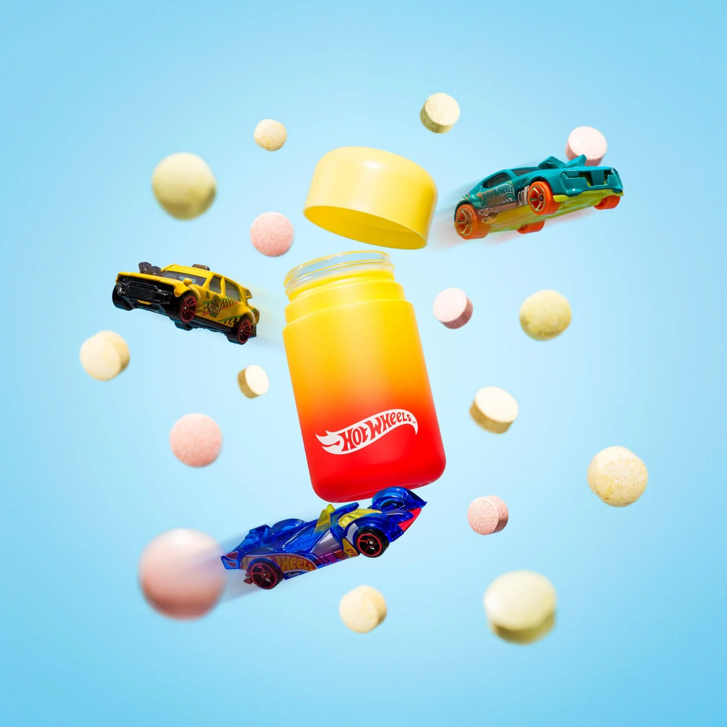 Hot Wheels container in yellow to orange gradient with yellow lid, surrounded by toy cars and colorful round candies or pills on a blue background.