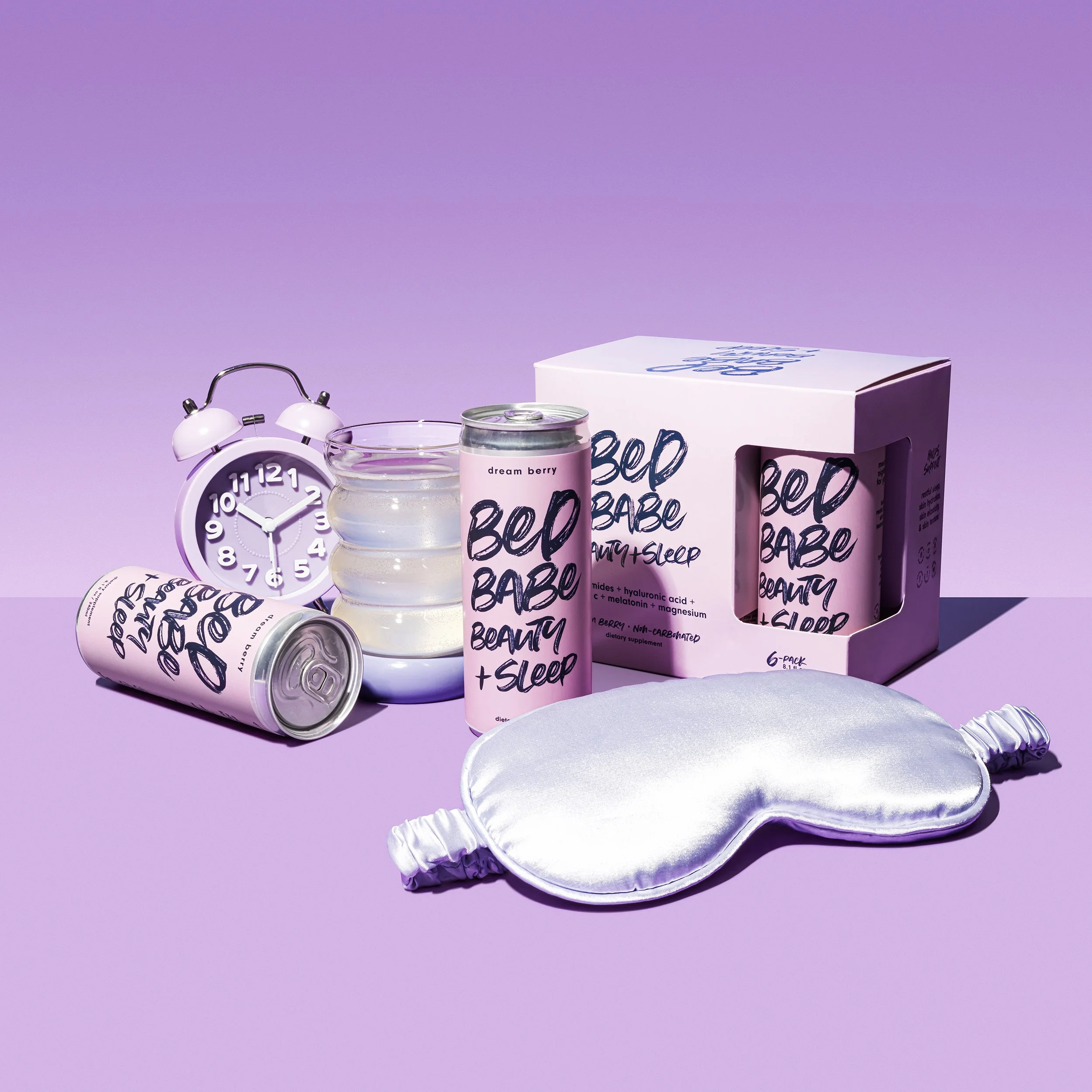 Pink cans labeled 'Bed Babe Beauty + Sleep,' a white box with similar labeling, a silver sleep eye mask, a small white alarm clock, a clear glass container with water, and a stacked set of round glass dishes, all set against a lavender purple backgro