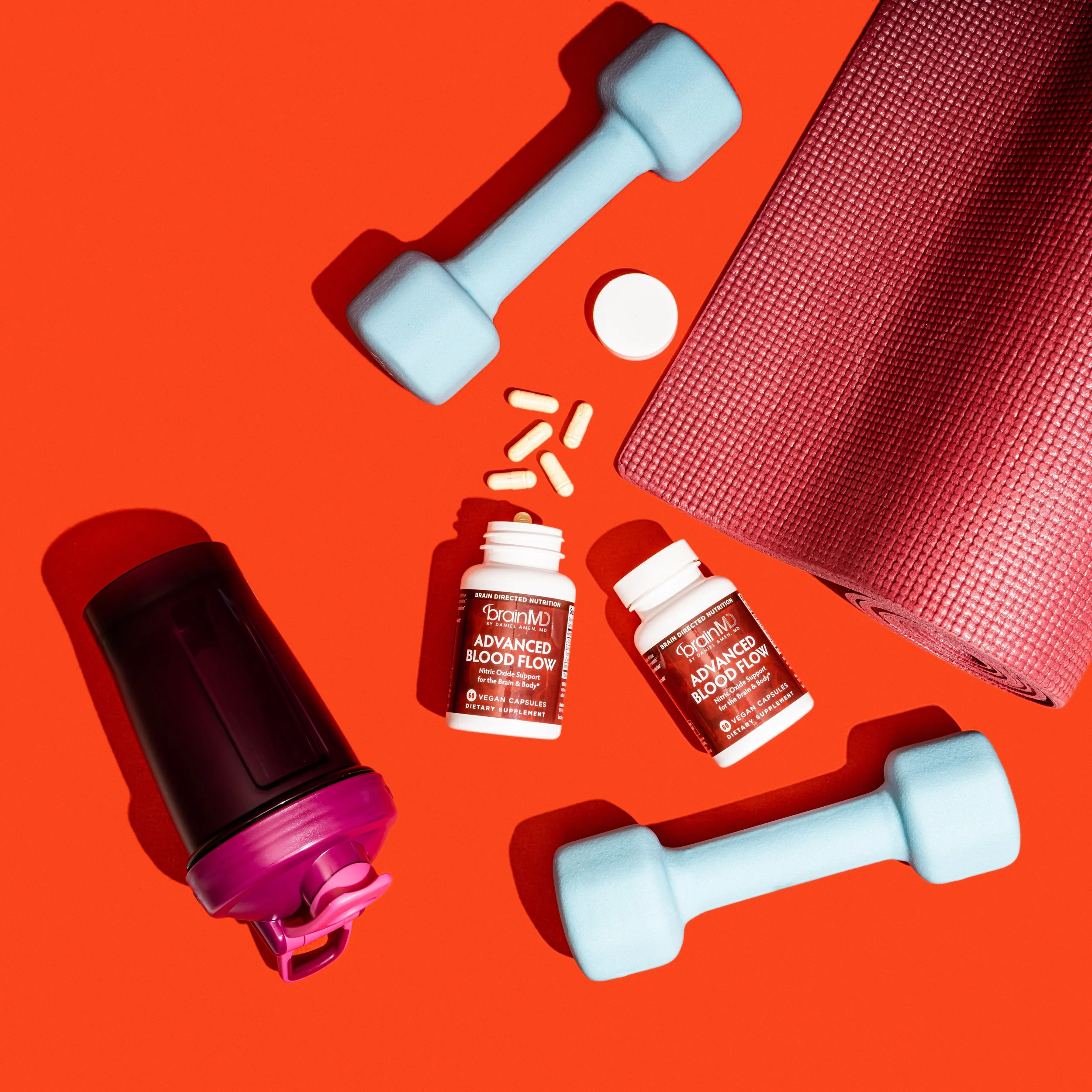 Dumbbells, pills, a water bottle, a rolled yoga mat, and dietary supplements on a red background.