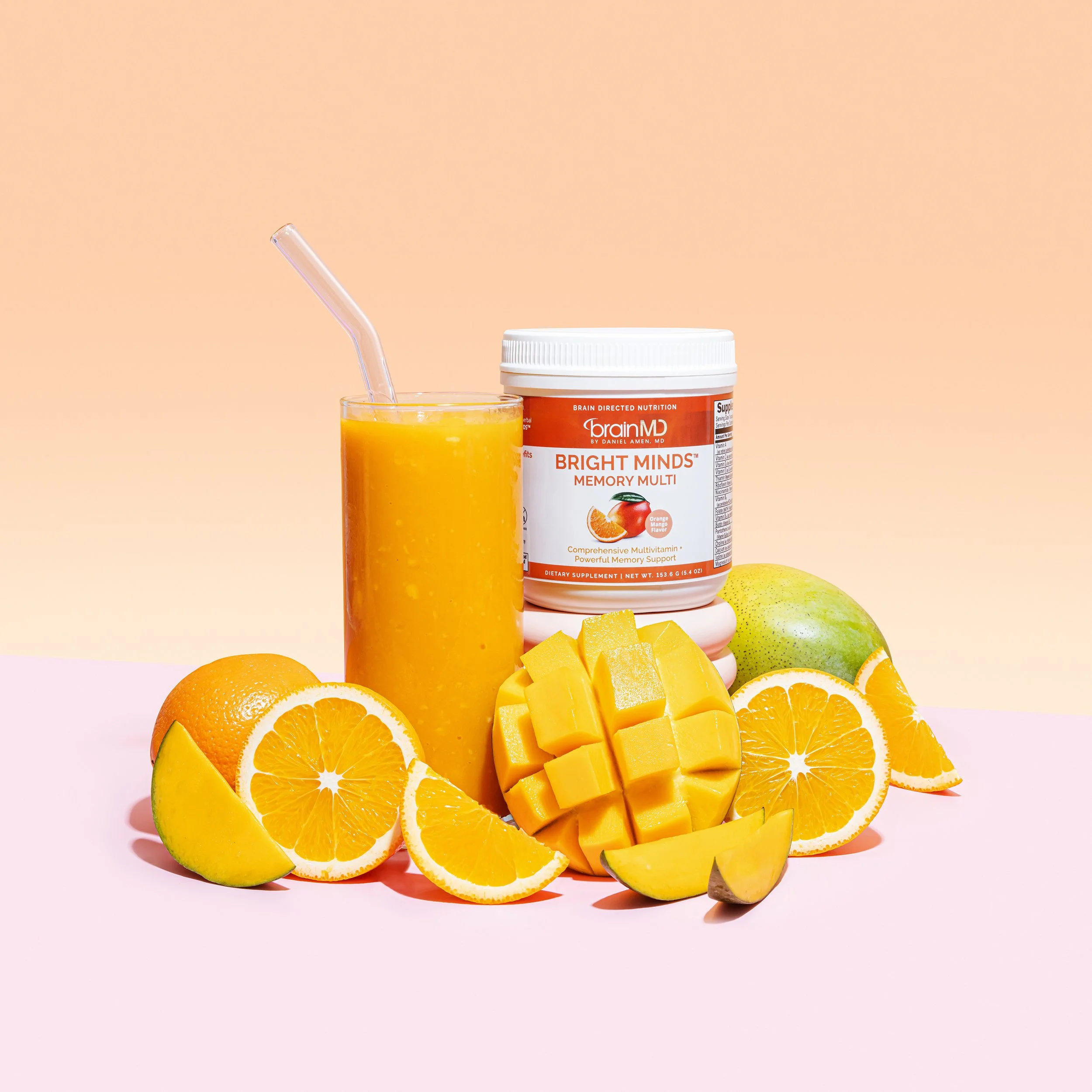 A glass of orange juice with a straw, a container of BrainMD Bright Minds Memory Multi supplement, and an arrangement of sliced oranges, mango, apple, and lemon on a pastel pink surface.