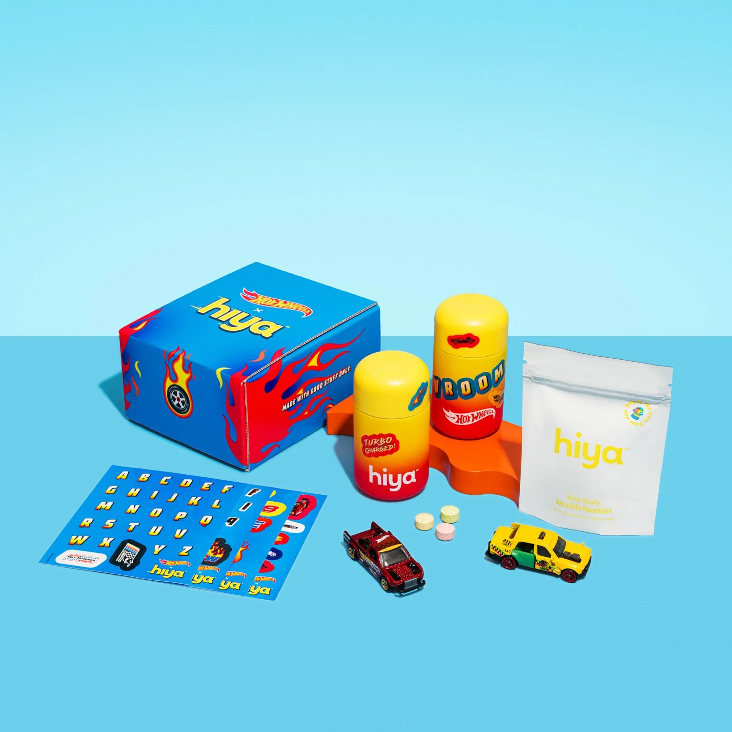 Hiya Kids Daily Multivitamin package, two yellow Toy Hot Wheels cars, two bottles labeled 'Turbo Charged' and 'Vroom', colorful alphabet stickers, and small tablets on a blue surface.