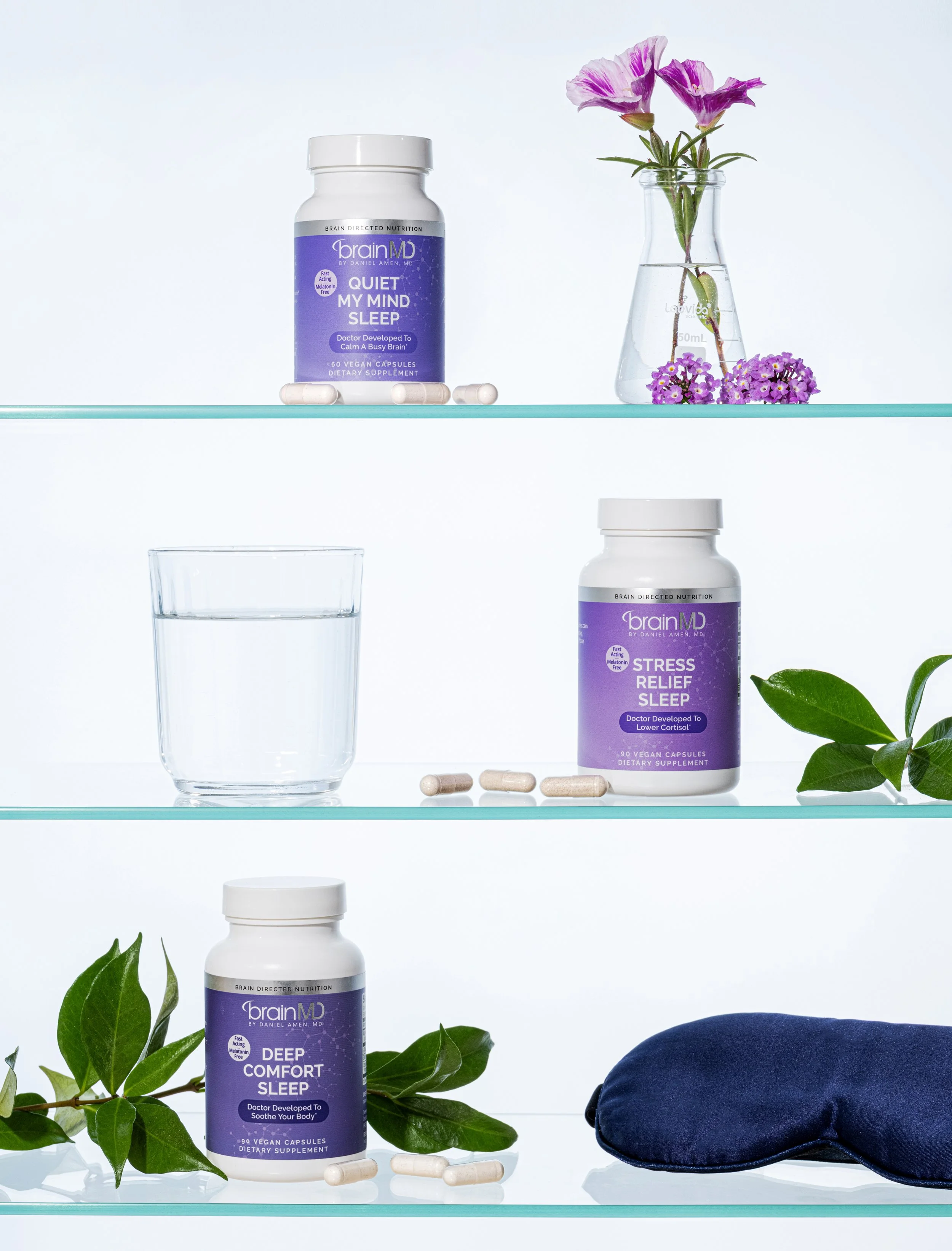 Three shelves with bottles of sleep aid supplements, a glass of water, a flower in a vase, green leaves, a navy sleep mask, and scattered capsules.
