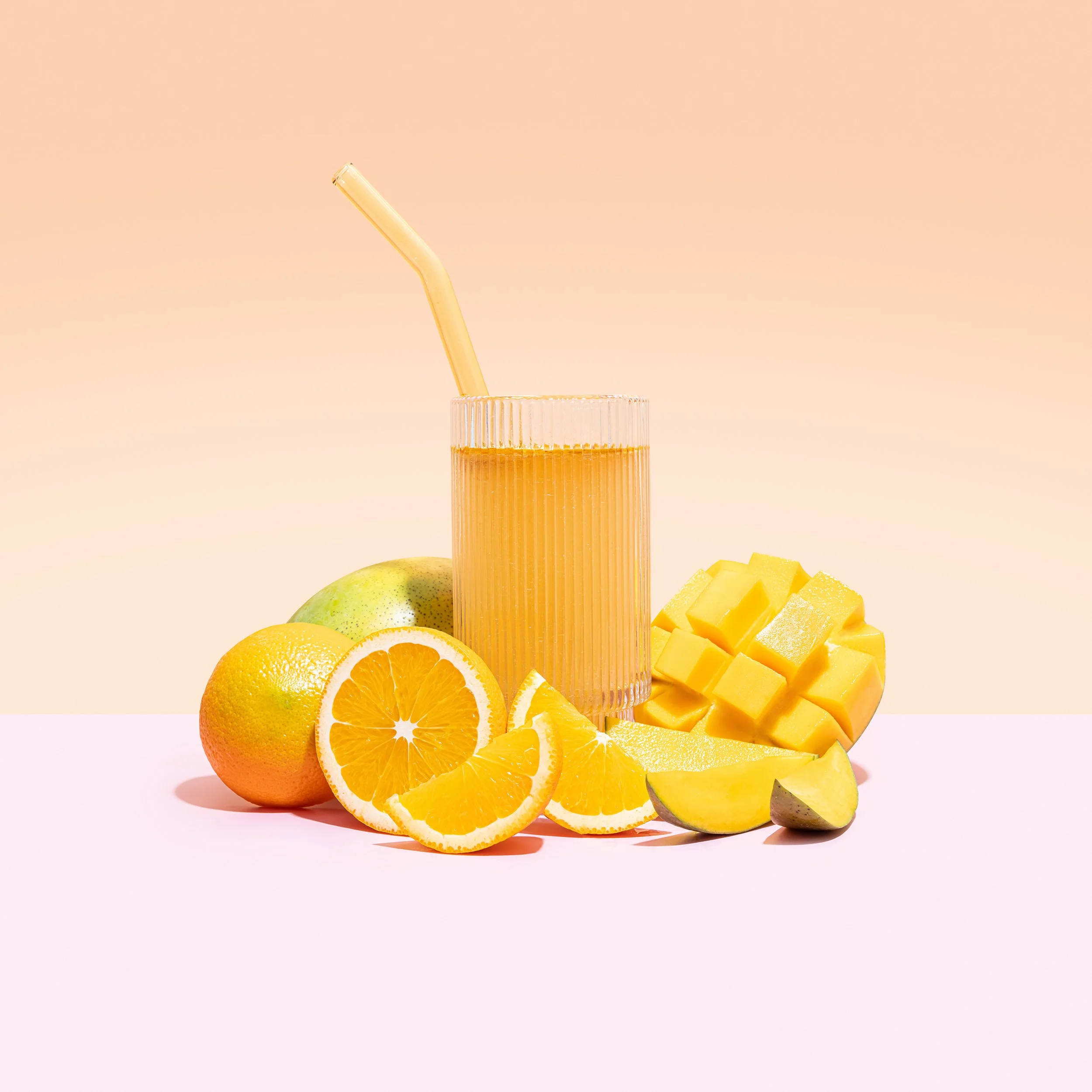 Glass of orange juice surrounded by oranges, mango, and slices of citrus on pastel pink and peach background.