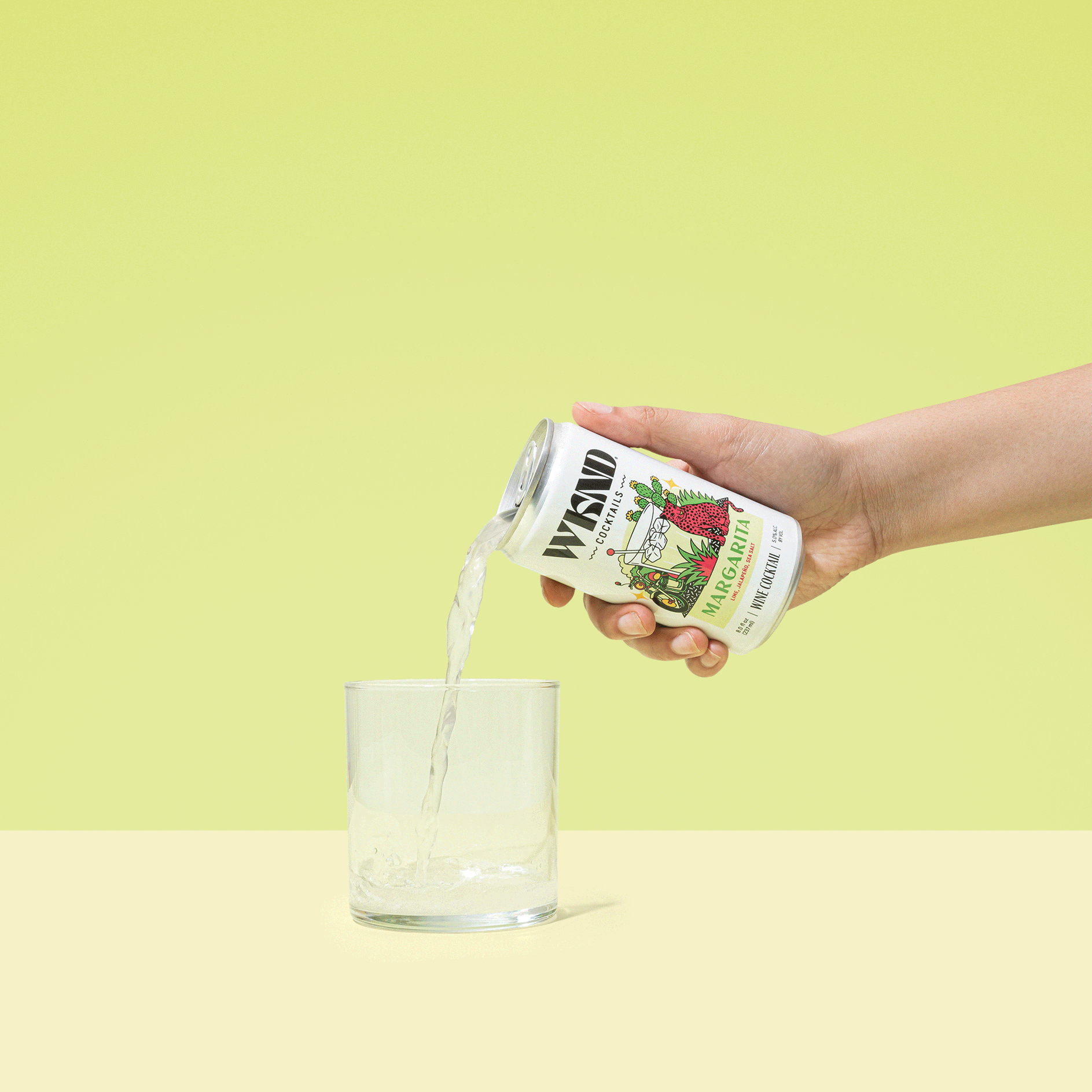 A hand pouring Wydad margarita canned cocktail into a glass against a pastel yellow and green background.