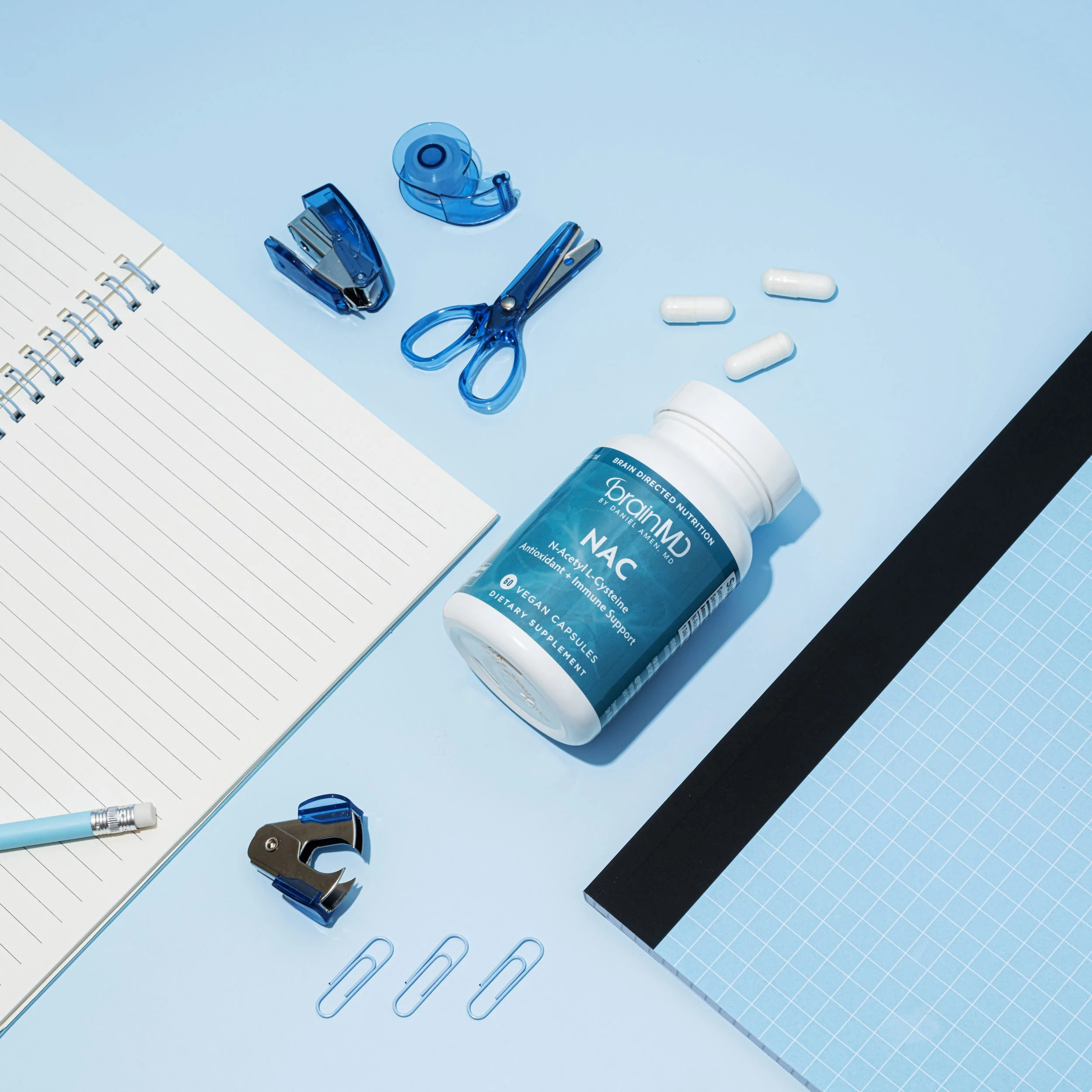 Blue stationery and supplement bottle on light blue background, including scissors, tape dispenser, paper clips, notebook, pen, and pills.