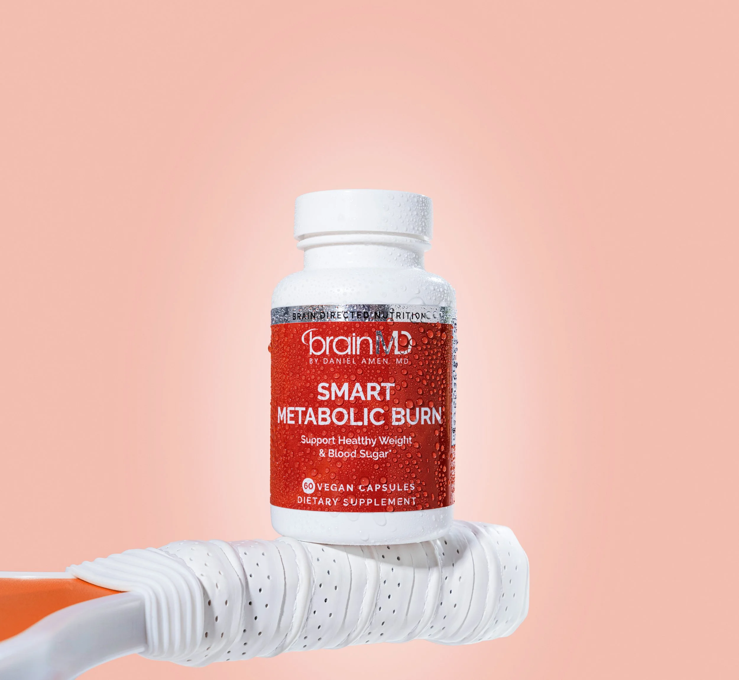 A white bottle labeled "Brain Mp SMART METABOLIC BURN" dietary supplement, with bubbles on the label, placed on a white textured surface against a pink background.