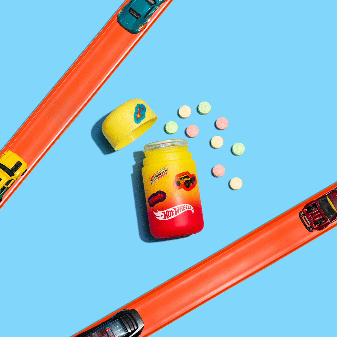 A Hot Wheels themed candy jar with colorful round candies spilling out, set on a blue background with orange toy racing tracks and two toy cars.