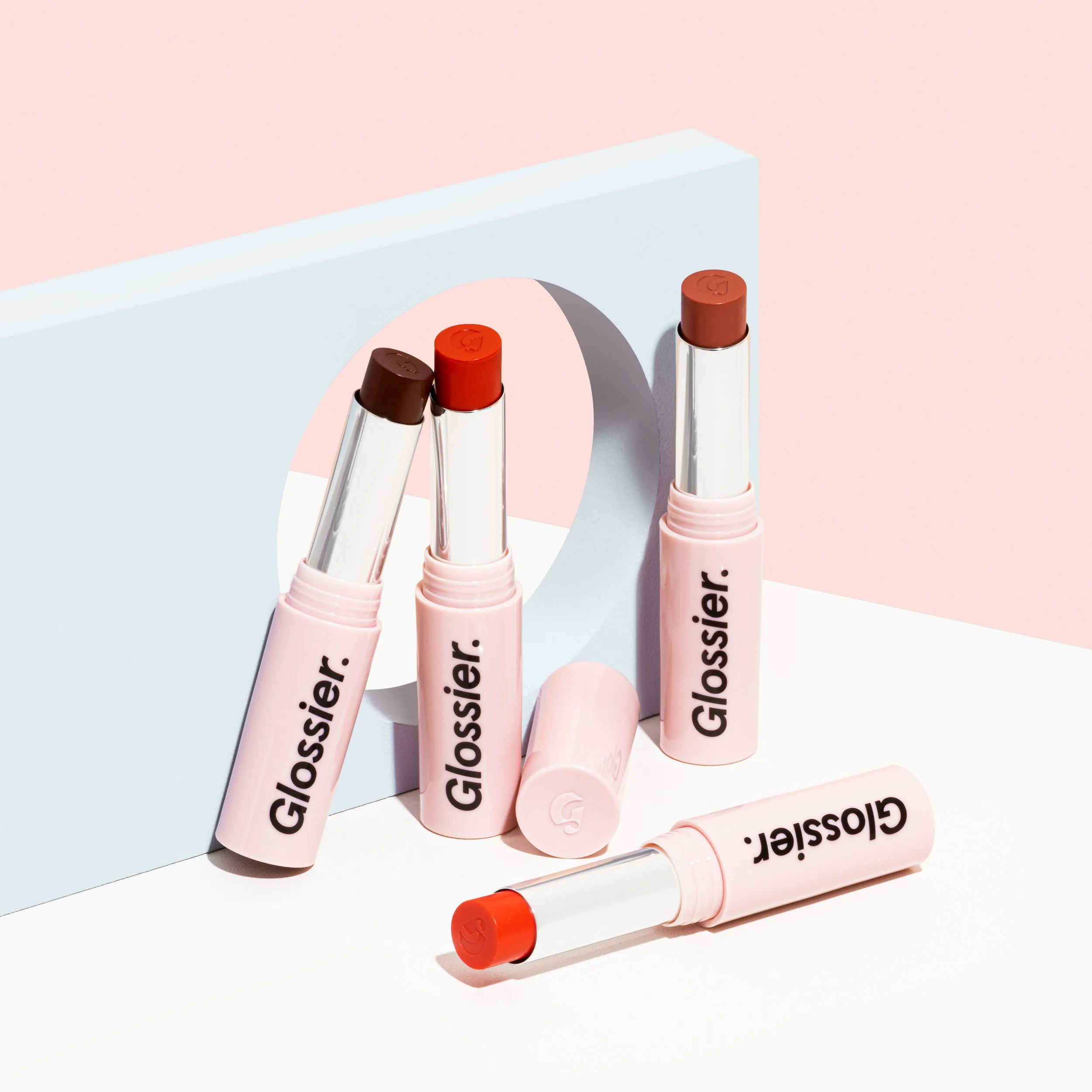 Four Glossier Lipstick Crayons, two with caps off revealing shades of brown and nude, against a pastel background with a large letter 'O' cutout.