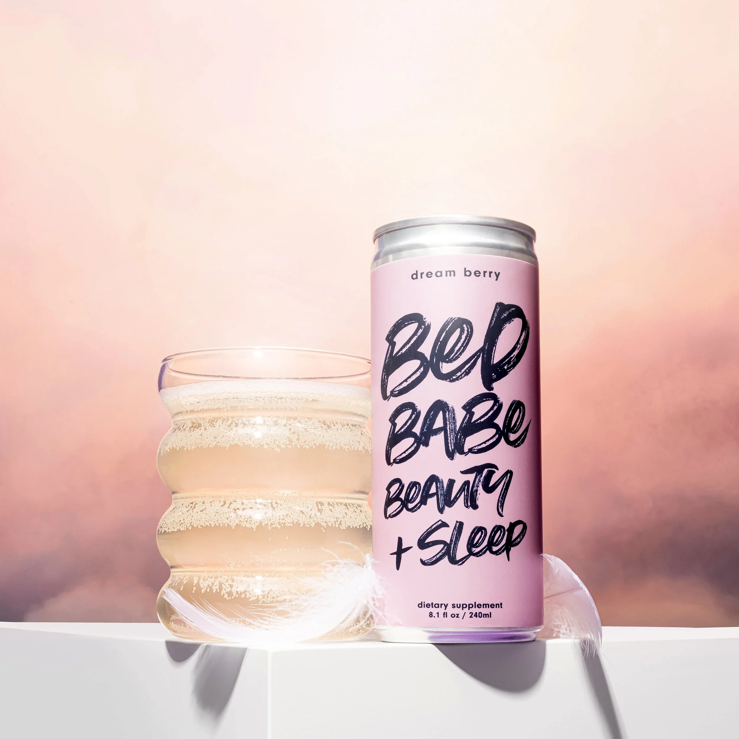 A can of Bed Babe Beauty + Sleep dietary supplement with pink design, placed next to a glass of creamy beverage, set against a soft pink and purple background.