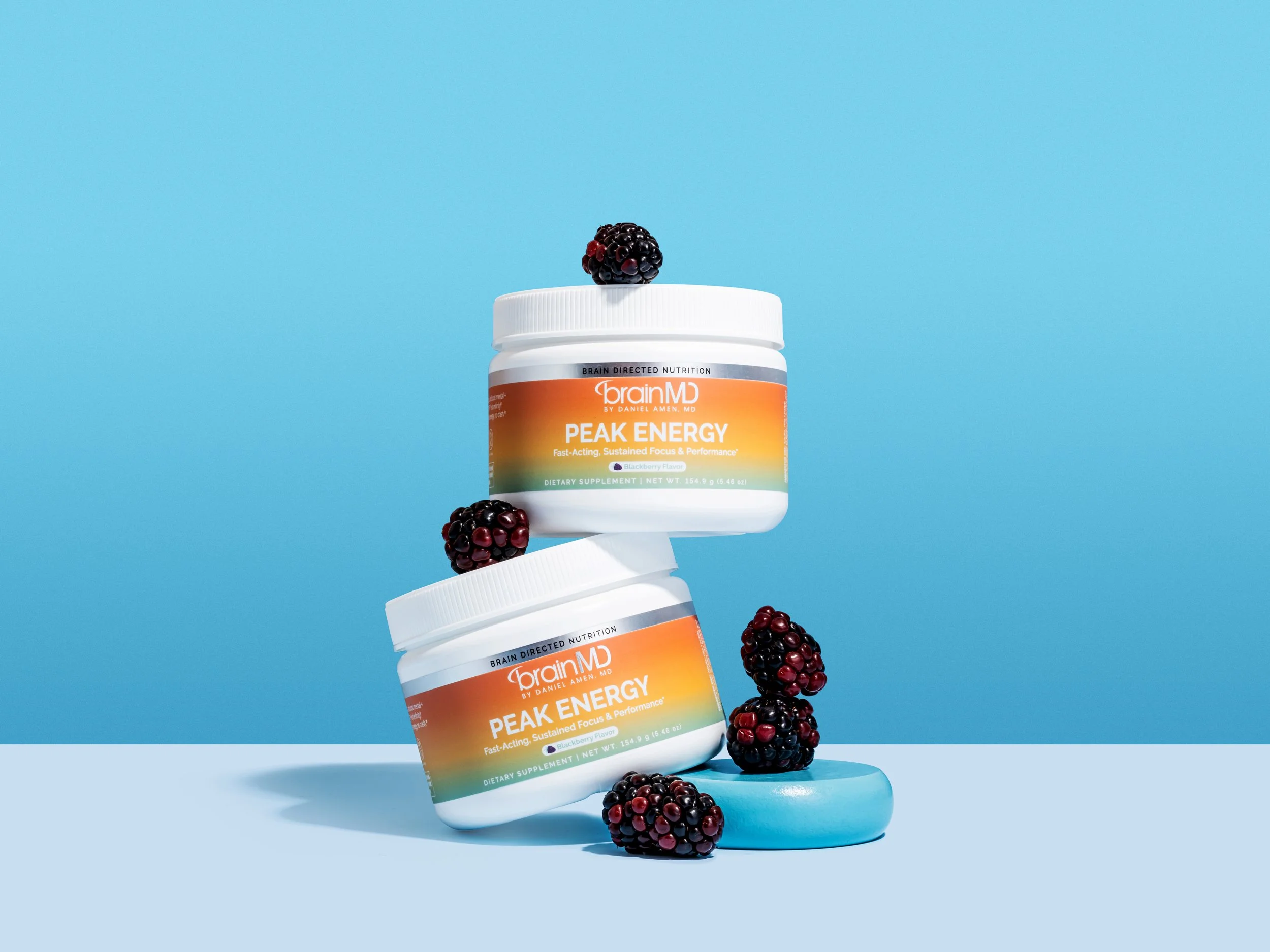 Two containers of BrainMD Peak Energy dietary supplements with blackberries and a blue object on a split blue surface against a blue background.