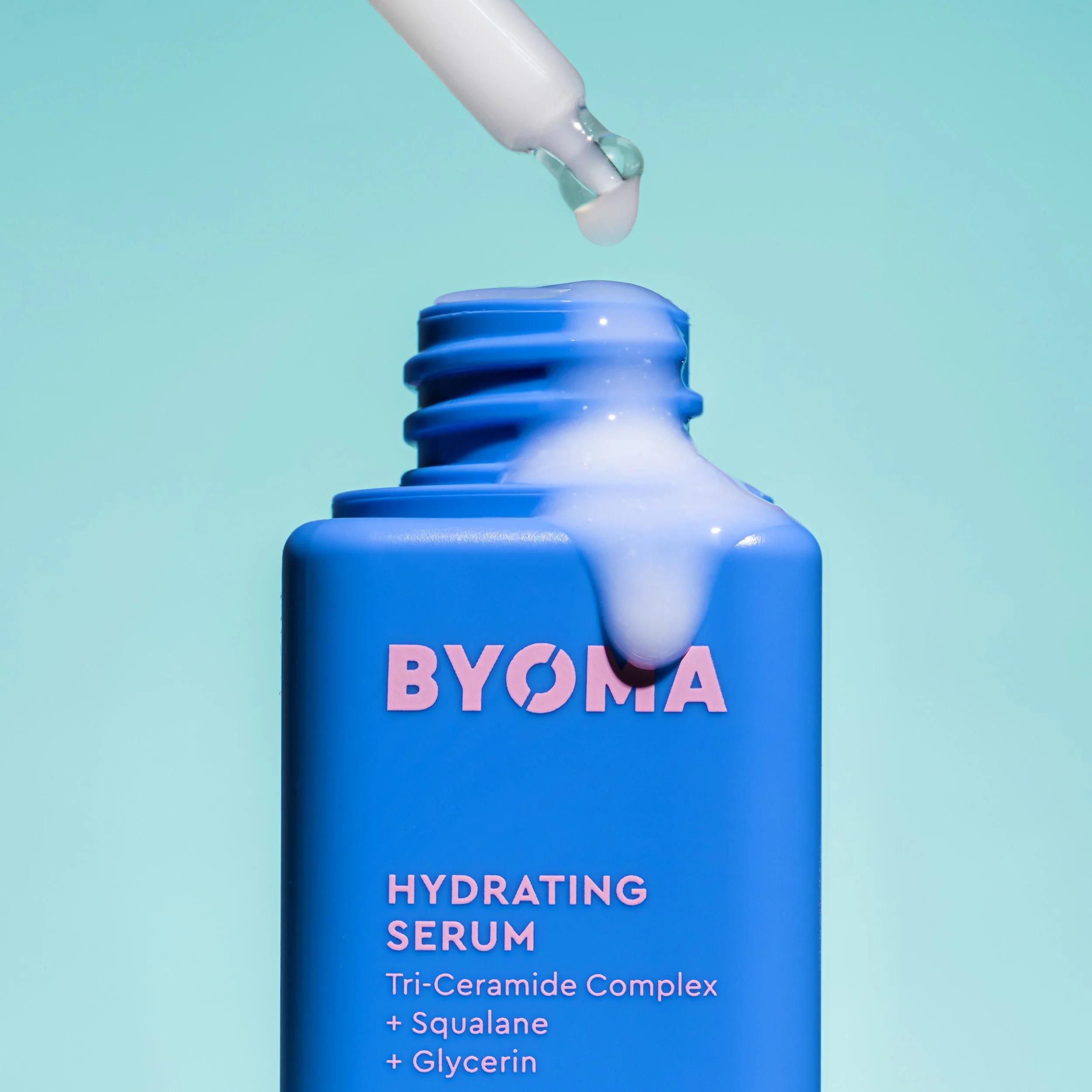 A blue bottle of BYOMA Hydrating Serum with a dropper releasing serum onto the opening of the bottle against a light blue background.