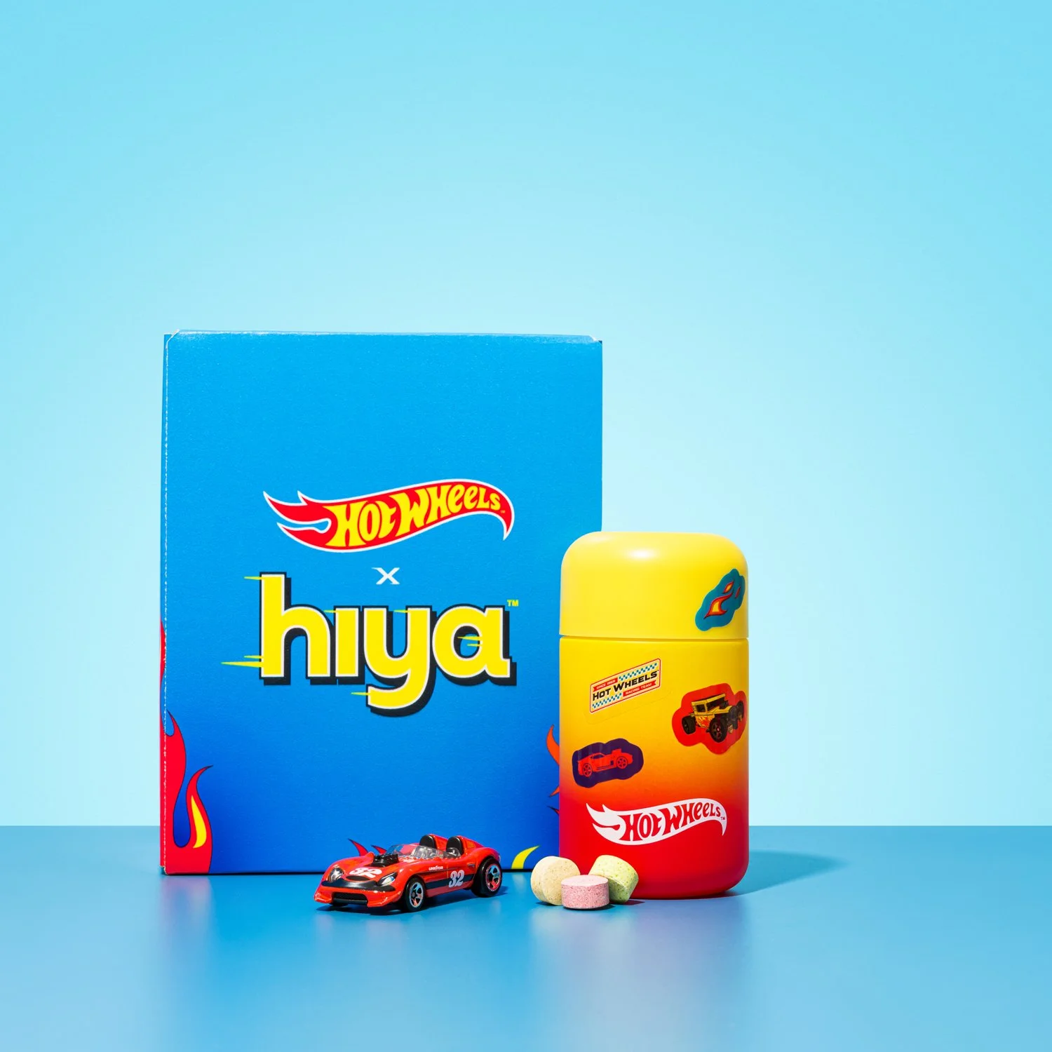 A hot wheels x hiya collaboration set with a blue box, a yellow container with hot wheels branding, a red toy car, and colorful pills on a blue surface with a light blue background.