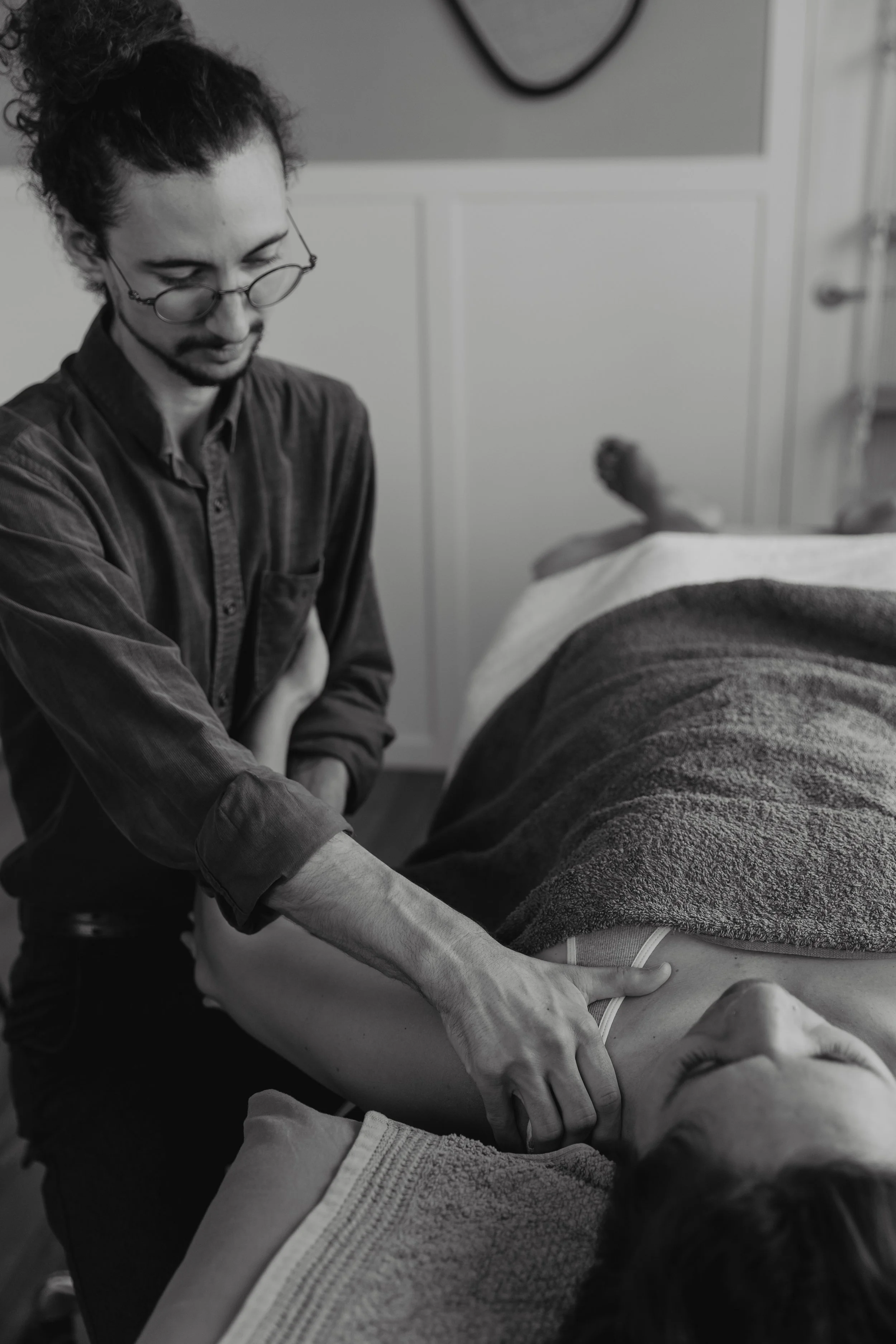 Kaspara Chaise, the osteopath, wearing glasses and a high bun, holding a patient's arm to move their shoulder while they are lying down in the treatment room.