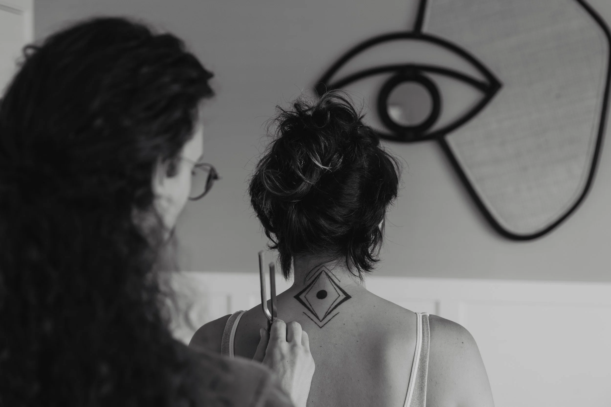 Kaspara Chaise demonstrating a type of sensory examination with a tuning fork on a pain patient's back. We can see a tattoo of a geometric design on the back of the patient's neck, with an abstract art piece on the wall in front of them.