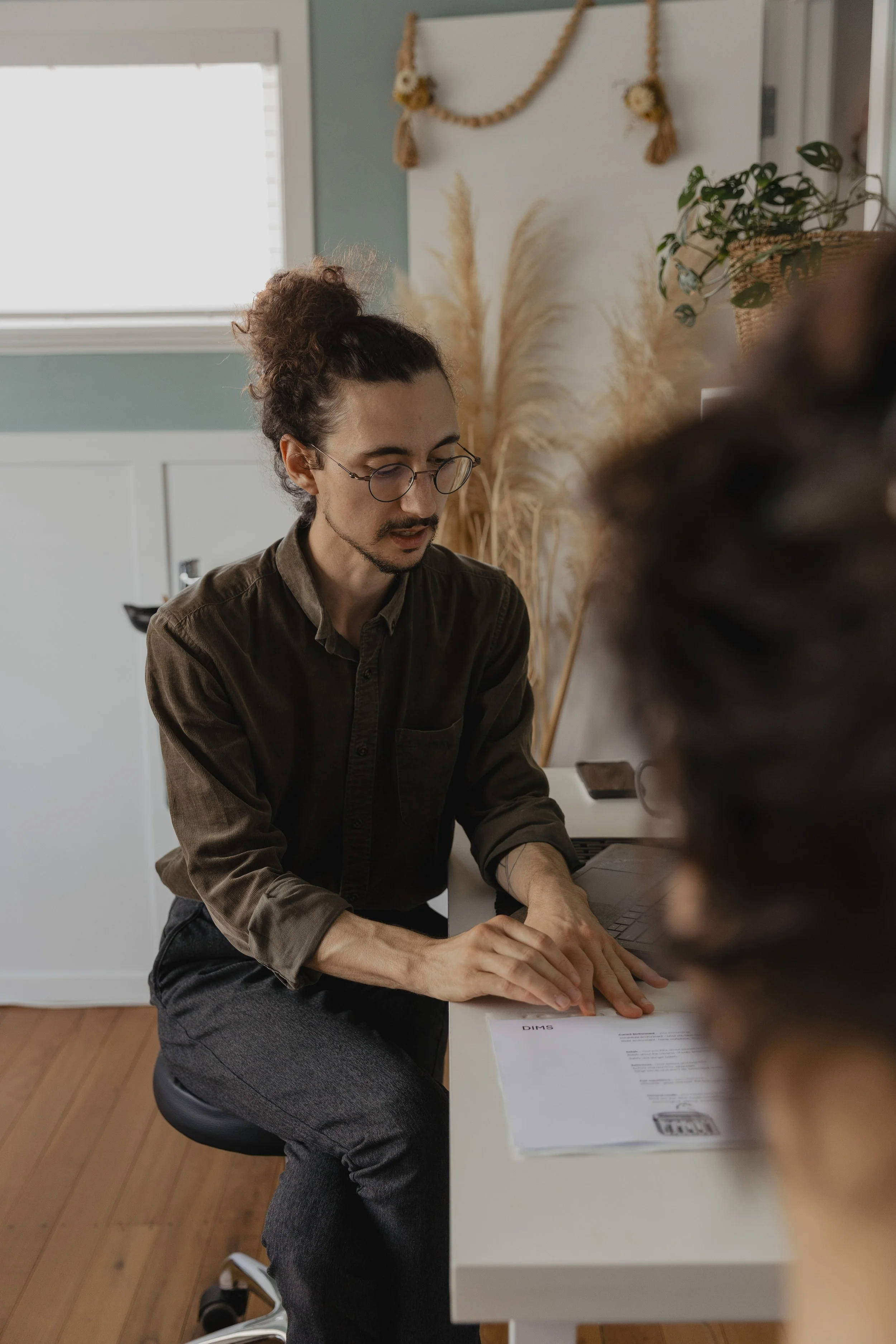 Kaspara Chaise, the osteopath, a man with long hair tied in a bun, glasses, and a beard, wearing a green shirt and gray pants, sitting at a white desk with a printed document explaining something to a patient at the chronic pain clinic.