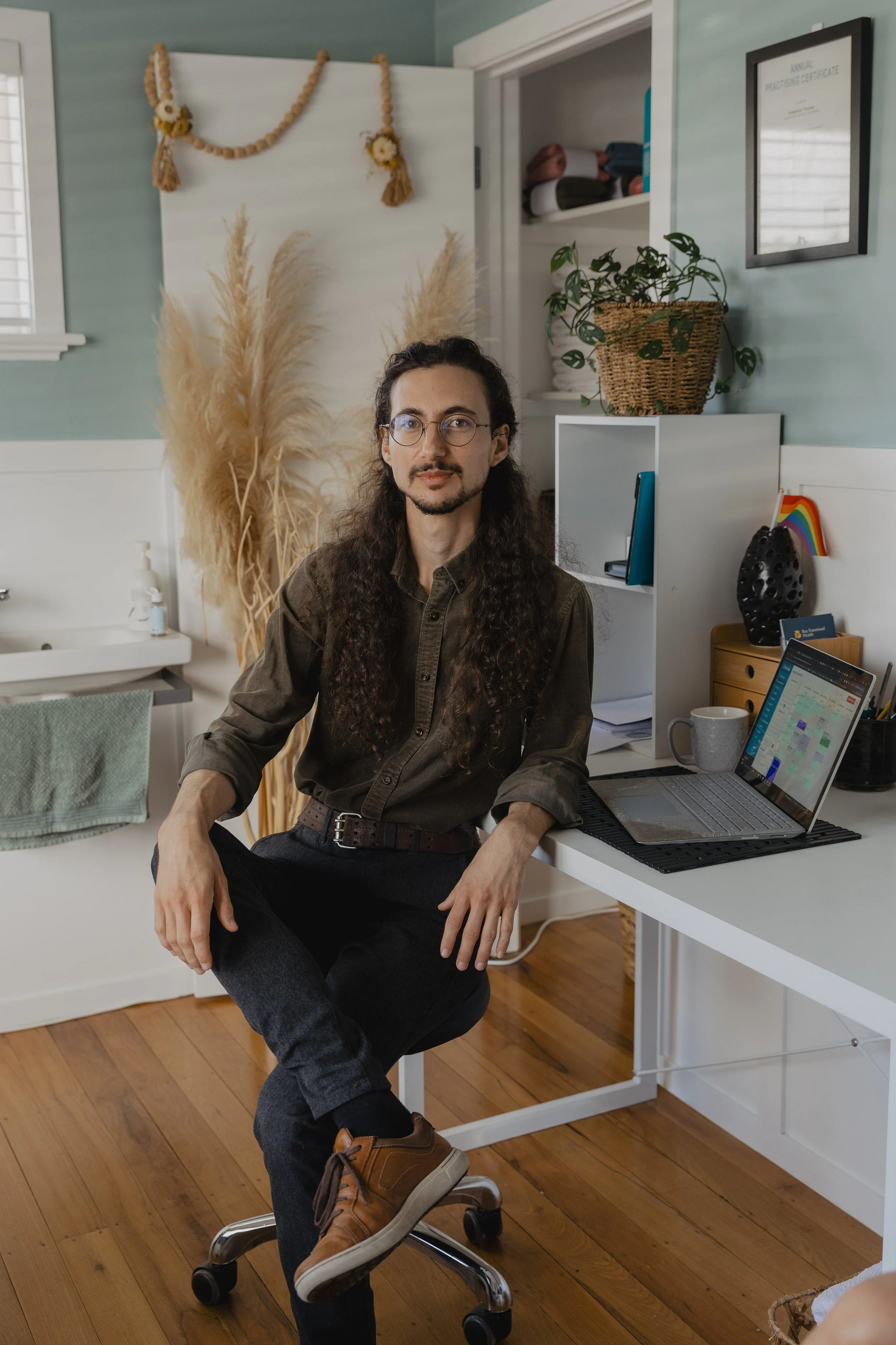 Kaspara Chaise, the osteopath, sits at his desk in the Ifix Health Clinic. The room has light blue walls, with plants and natural decorations. The desk has a laptop, a mug, office supplies and a small rainbow flag in a corner.