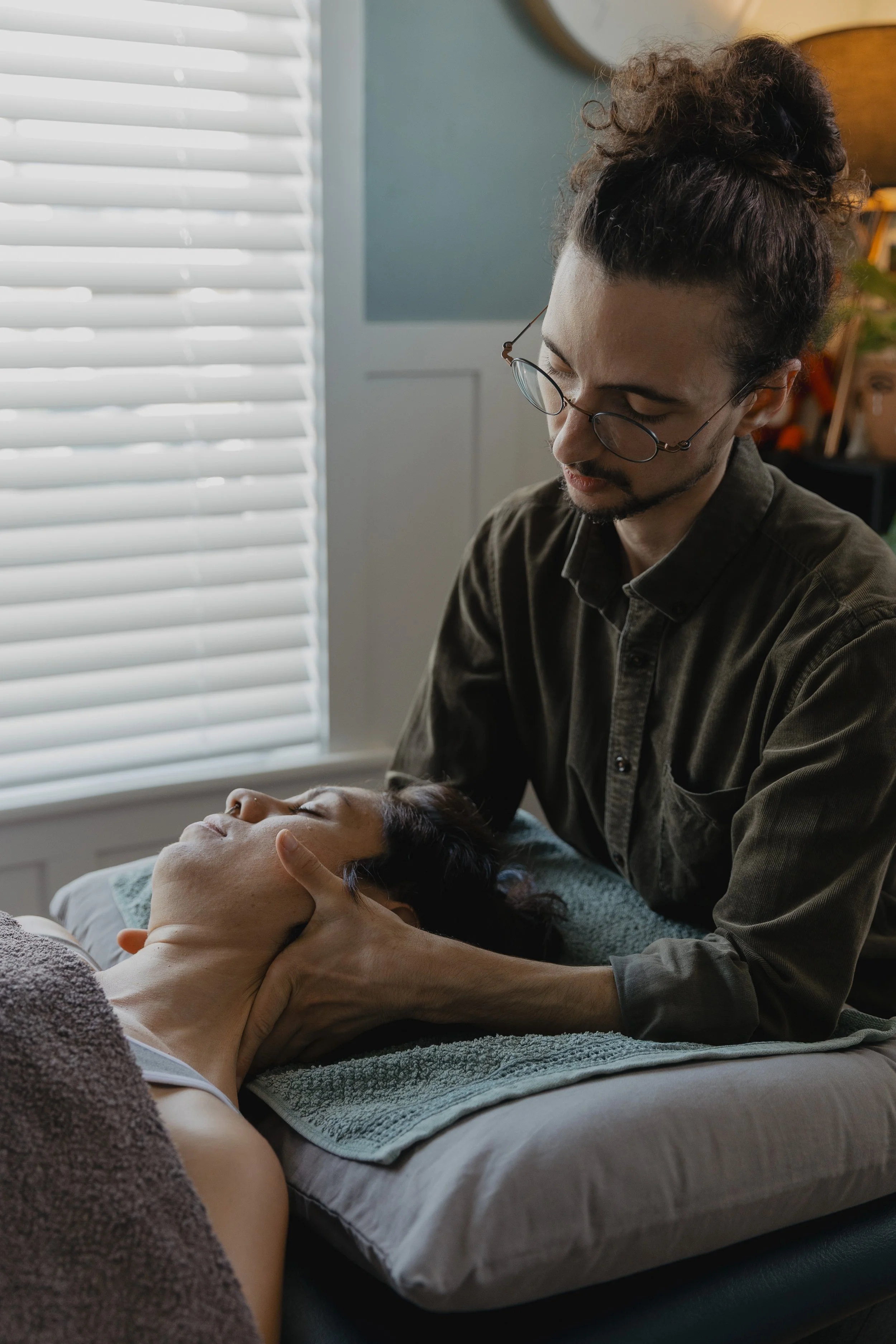 Kaspara Chaise expertly holds and moves the a neck of a patient lying on the treatment eyes closed in the treatment room with closed window blinds in the background.