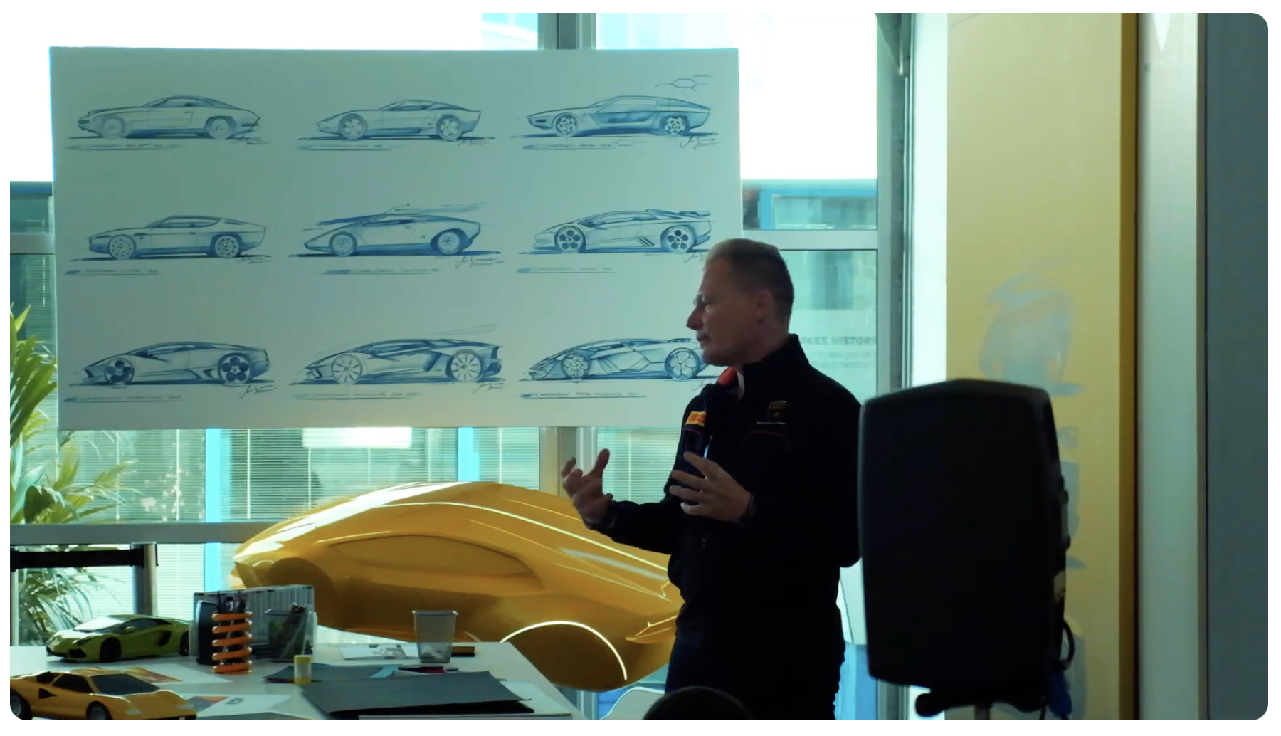 A man giving a presentation about car design, with sketches of sports cars on a large whiteboard behind him, and a yellow model car on a table in front.