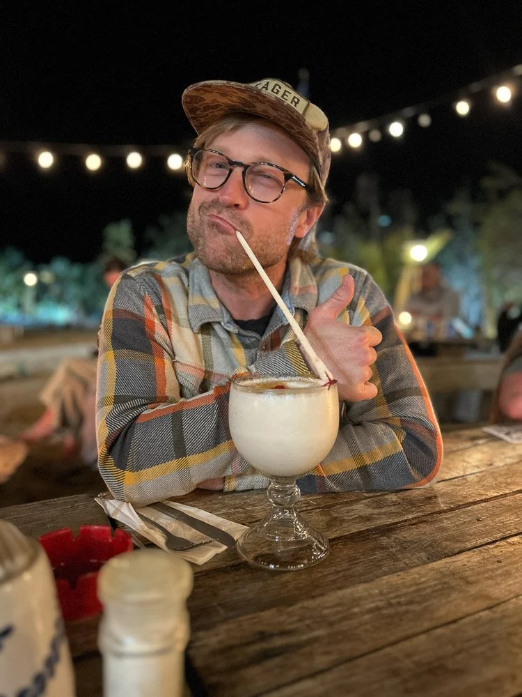 Man with glasses and hat sitting at a wooden table with a frosty drink, straw, and spoon, making a thumbs up gesture outdoors at night with string lights overhead.