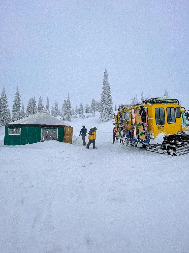 A snow-covered landscape with a yellow snowcat vehicle and a green tent, where several people are standing and preparing for outdoor snow activities, surrounded by snow-covered trees.