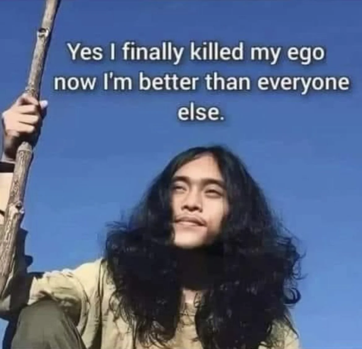 Person with long dark hair holding a stick against a blue sky, with overlaid text that reads, "Yes I finally killed my ego now I'm better than everyone else."