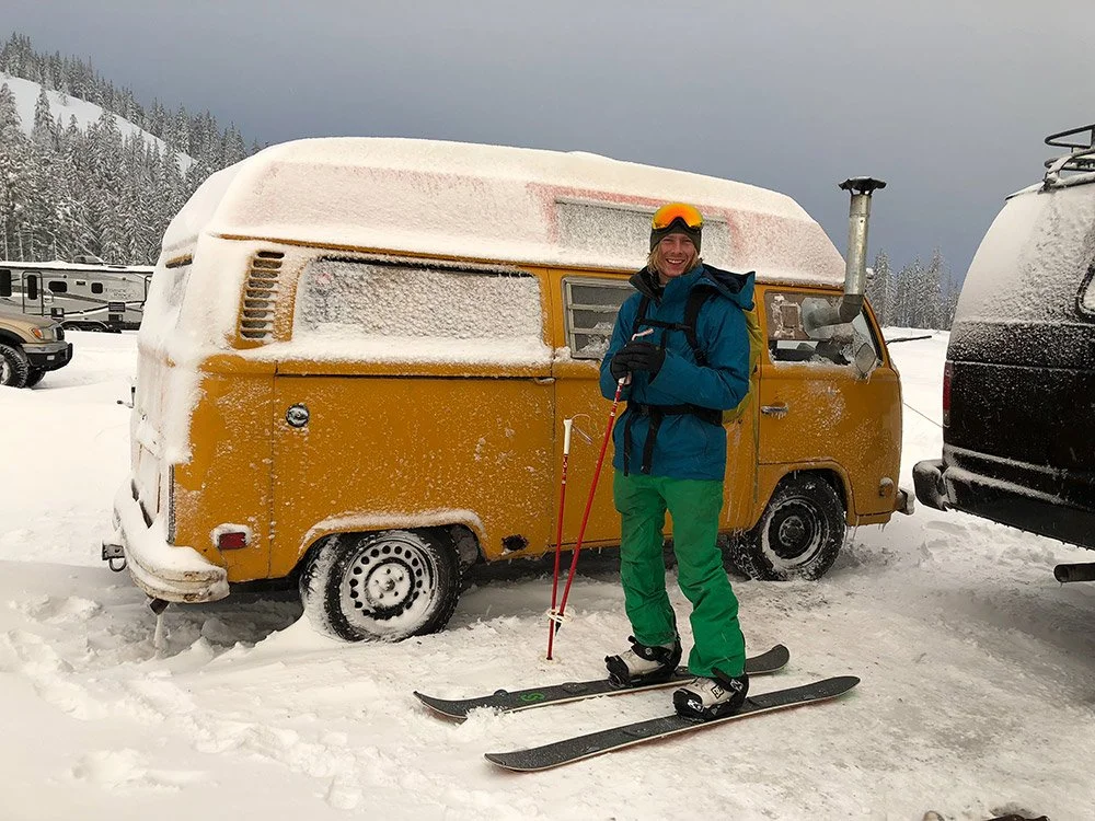 A person standing on skis in front of a yellow van covered in snow, holding ski poles and smiling, with snow-covered trees and vehicles in the background.