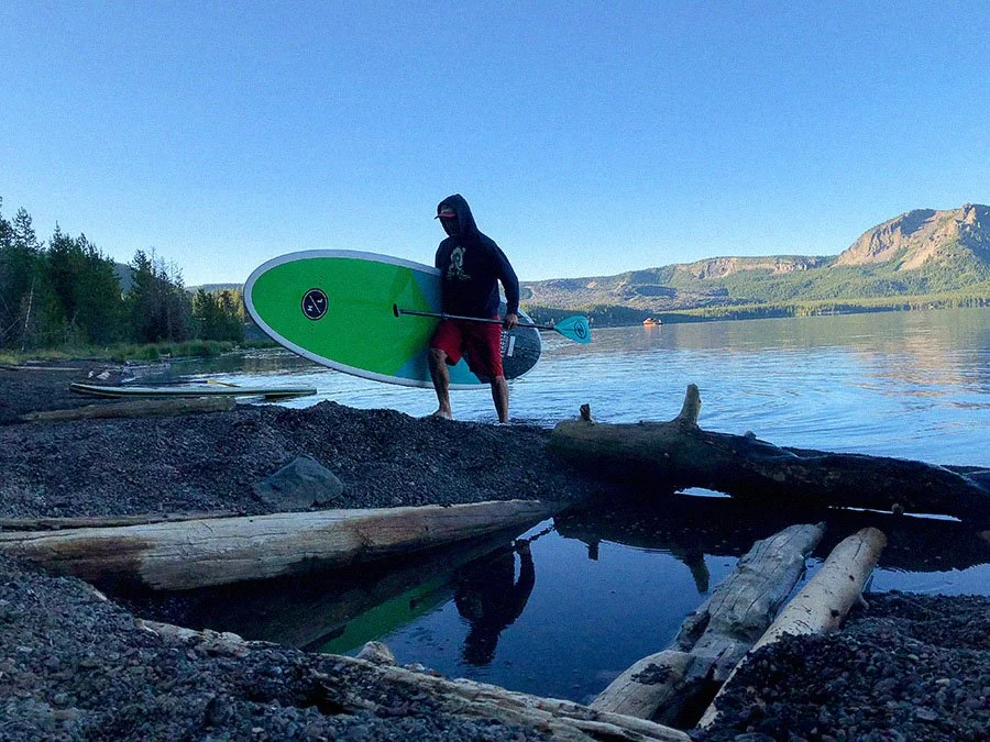 A person in a hooded jacket, shorts, and sunglasses holding a green paddleboard with a paddle, standing on a rocky shoreline by a calm lake with mountains in the background.