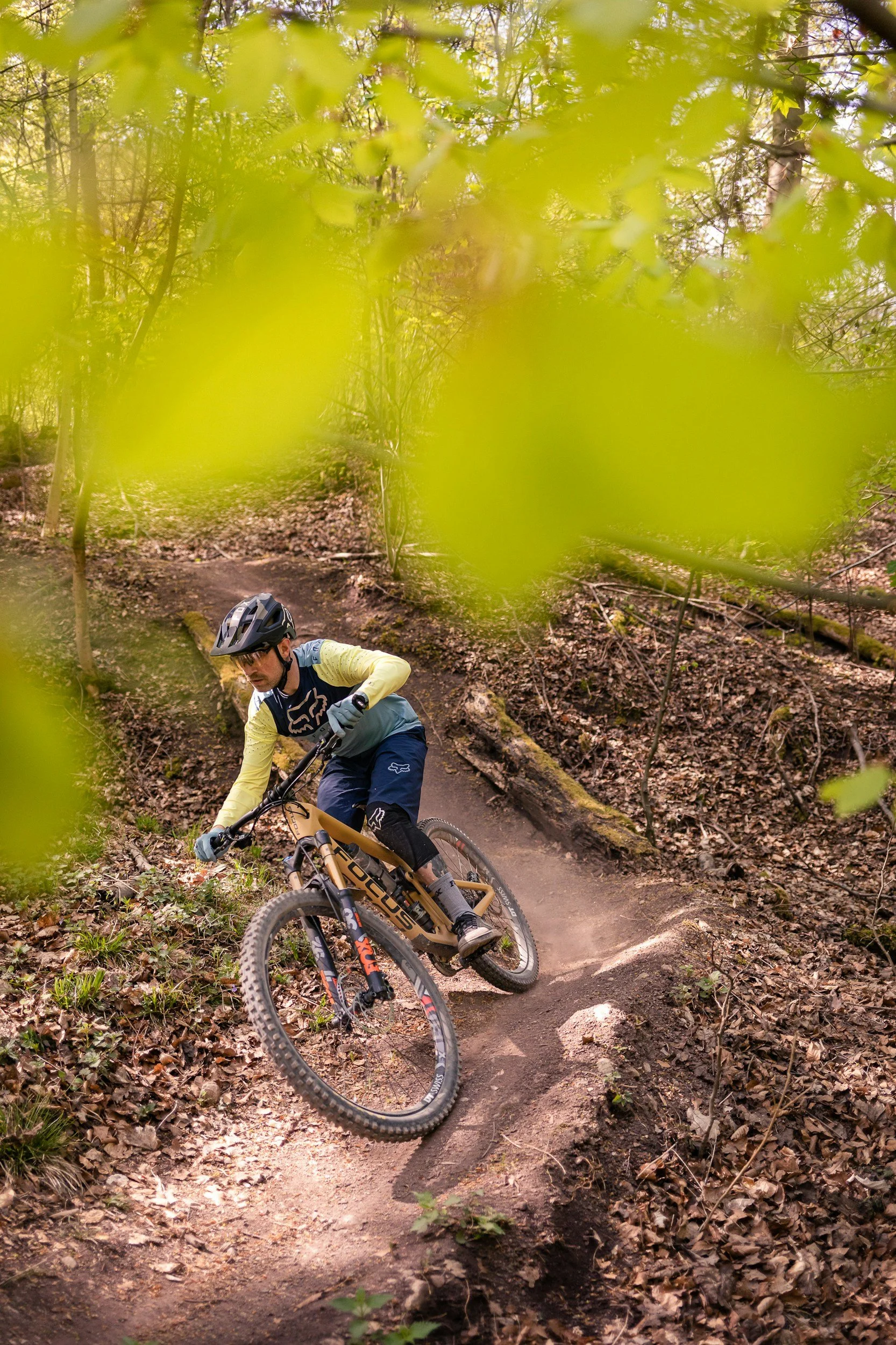 A mountain biker wearing a helmet and gloves navigates a winding trail through a wooded forest. The trail is narrow with dirt and leaves, surrounded by green foliage and trees.