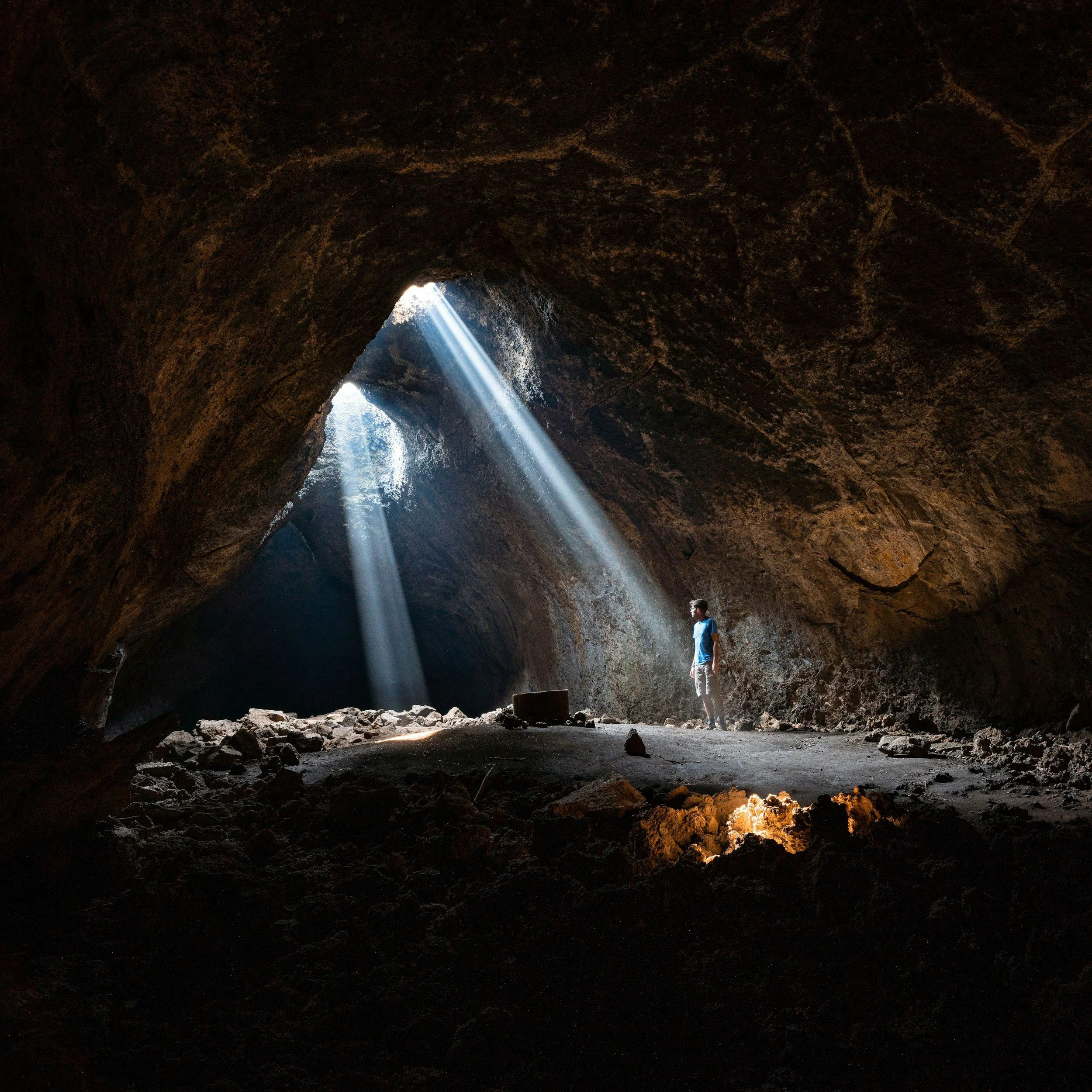 A person inside a dark cave with sunlight streaming in through an opening in the ceiling, illuminating the rocky floor and casting beams of light.
