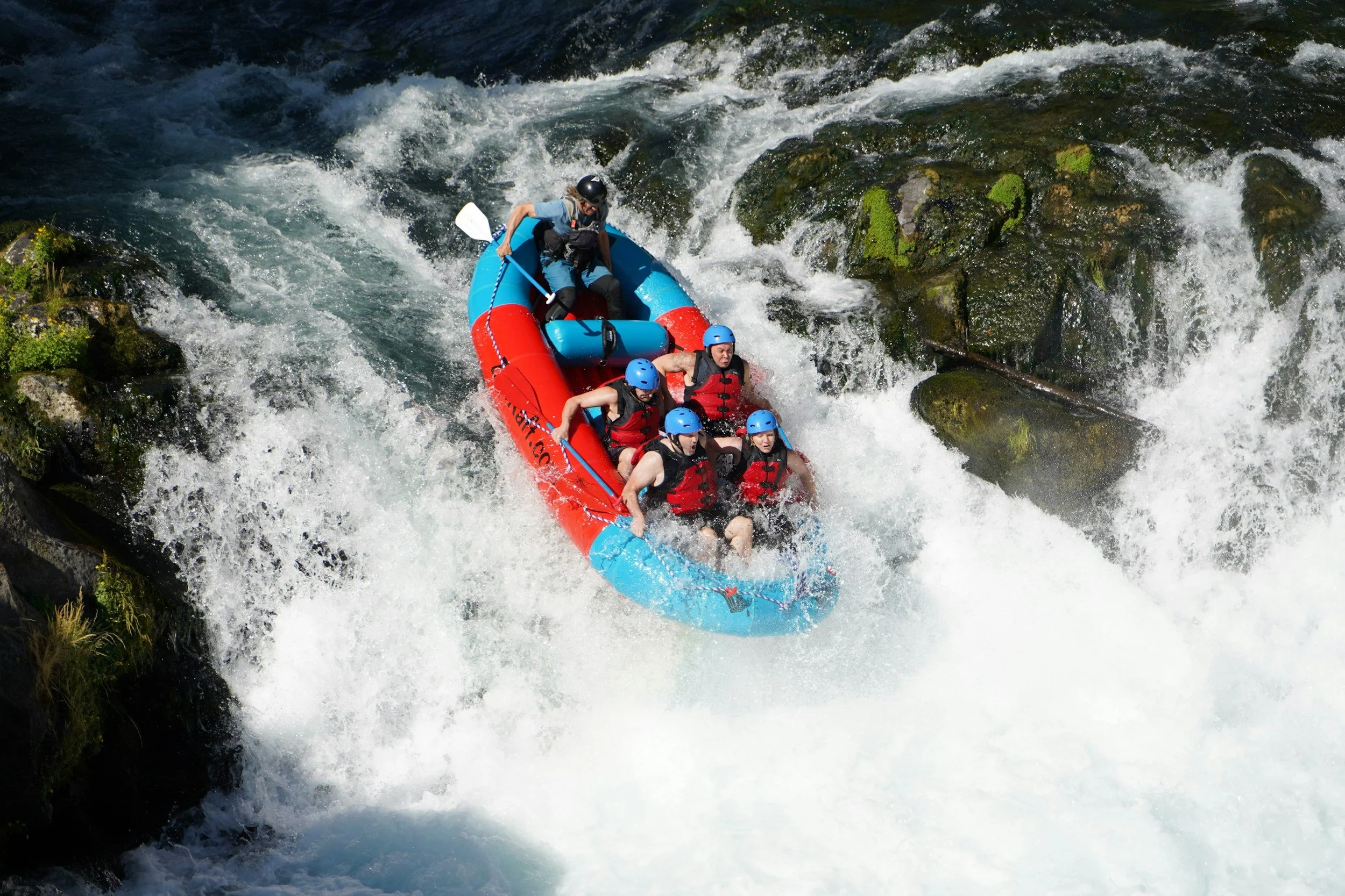 A group of people white water rafting on a river, wearing life jackets and helmets, navigating through rapids with rocks and water splashing around.