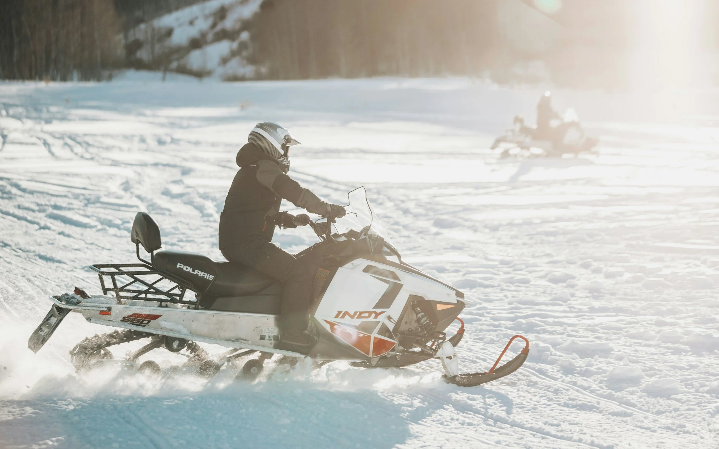Person riding a Polaris snowmobile on snowy terrain with another snowmobile in the background during daylight.