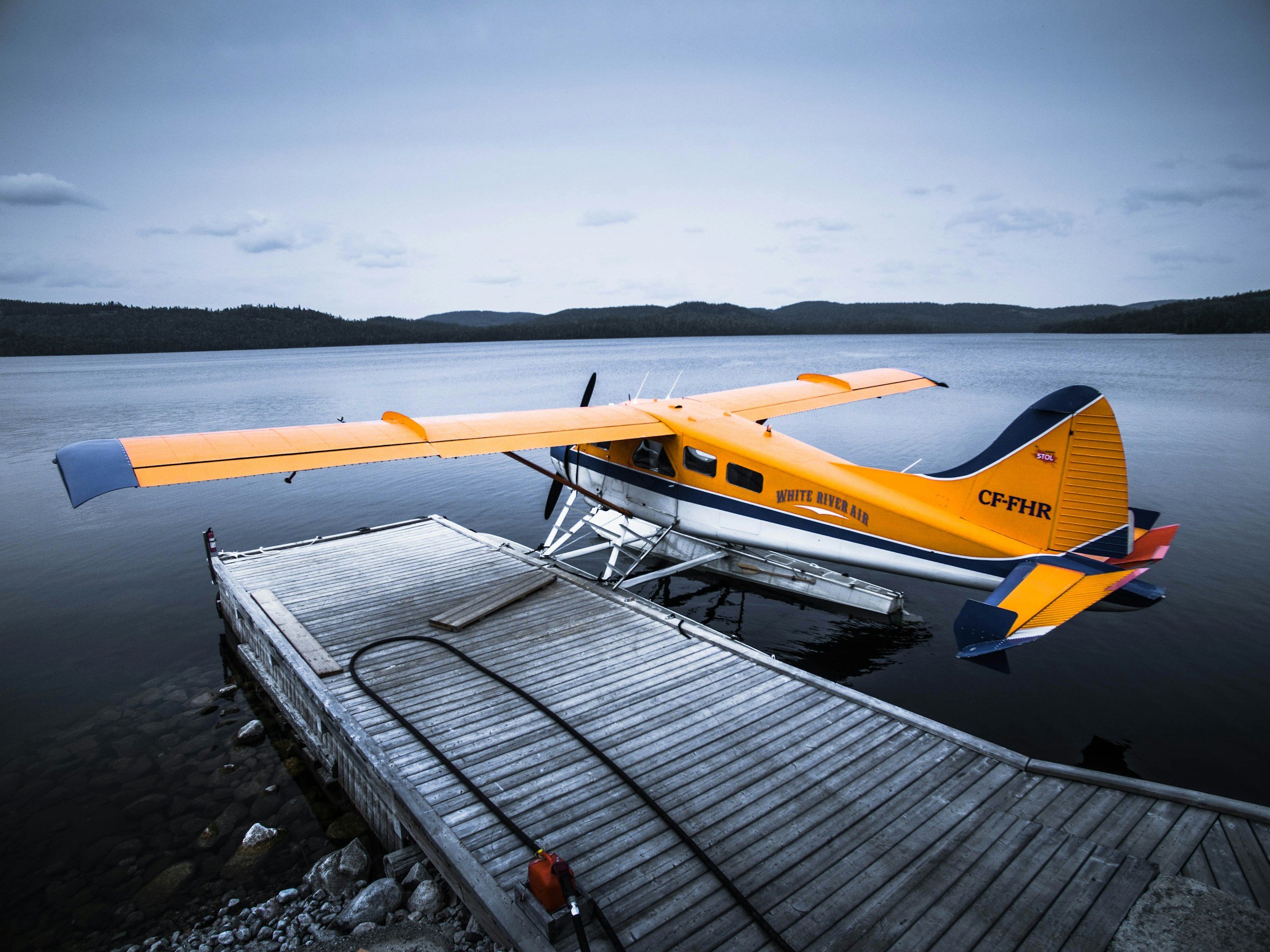 A yellow and white floatplane on a dock by a calm lake with wooded hills in the background.