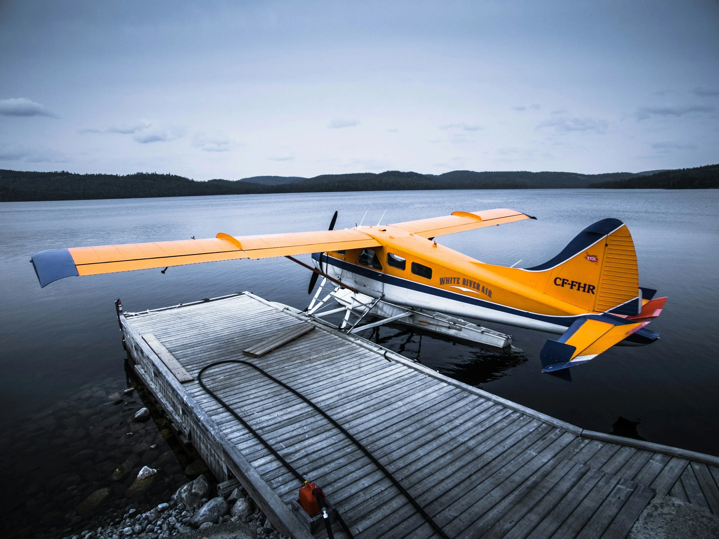 A small yellow floatplane on a dock extending into a calm lake, with forested hills in the background.