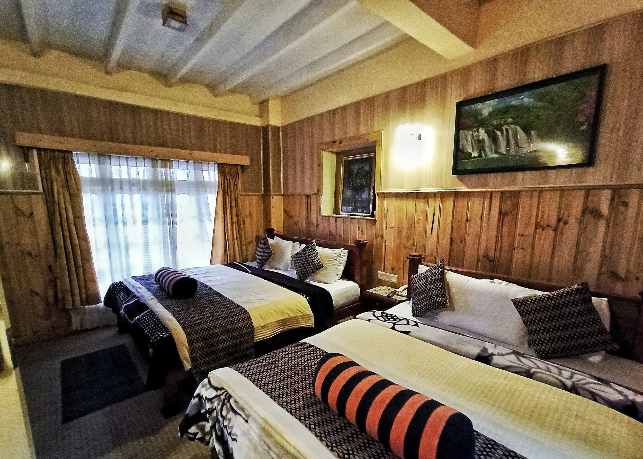 A cozy hotel room with wood-paneled walls, two beds with patterned pillows and throws, a window with curtains, a wall-mounted television, and a small window above one bed.
