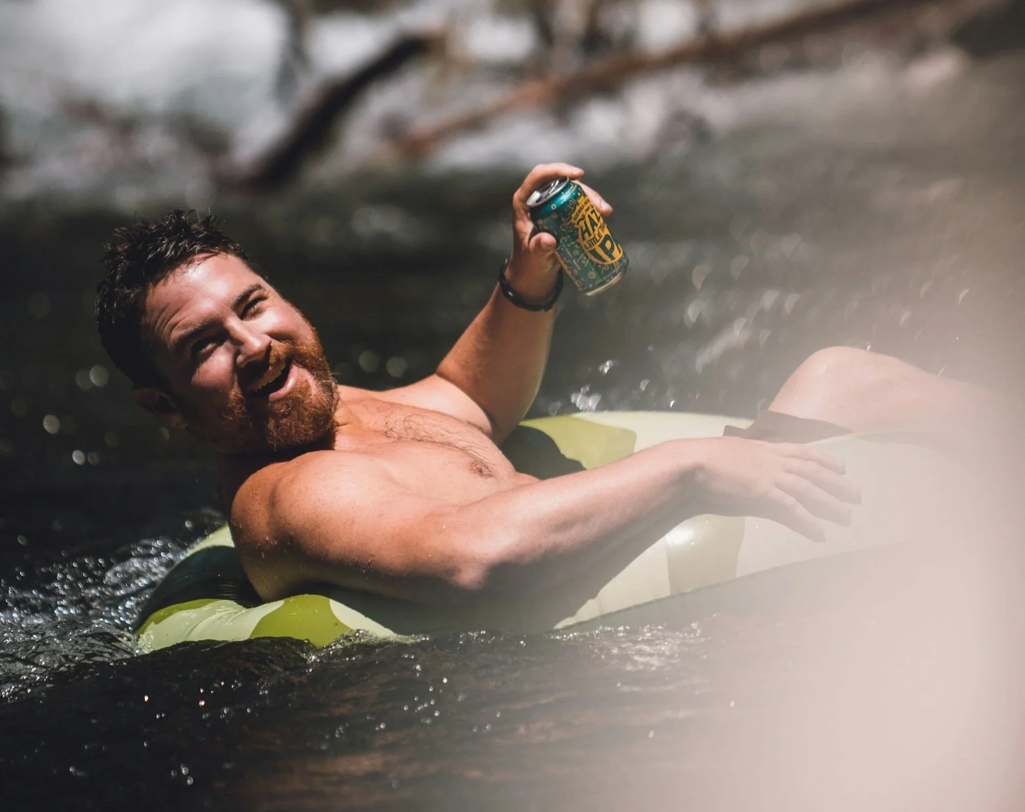 A man with a beard and mustache relaxing on an inflatable float in a river, holding a can of beer, smiling and enjoying the outdoor water activity.