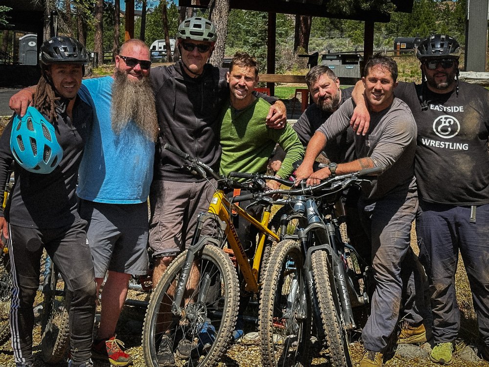 A group of seven men, some with beards and wearing helmets, standing outdoors with bicycles, in a wooded area.