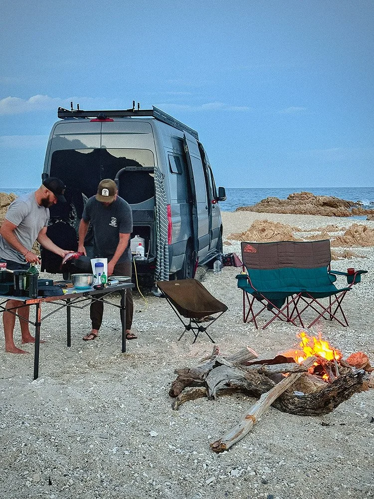 Two people preparing food on a camping table near a van on a rocky beach, with a campfire burning nearby, and folding chairs set up facing the ocean.