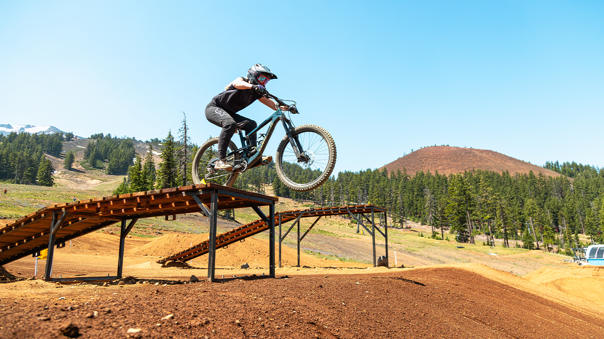 A person mountain biking jumps over a ramp on an outdoor dirt trail with green trees and hills in the background.