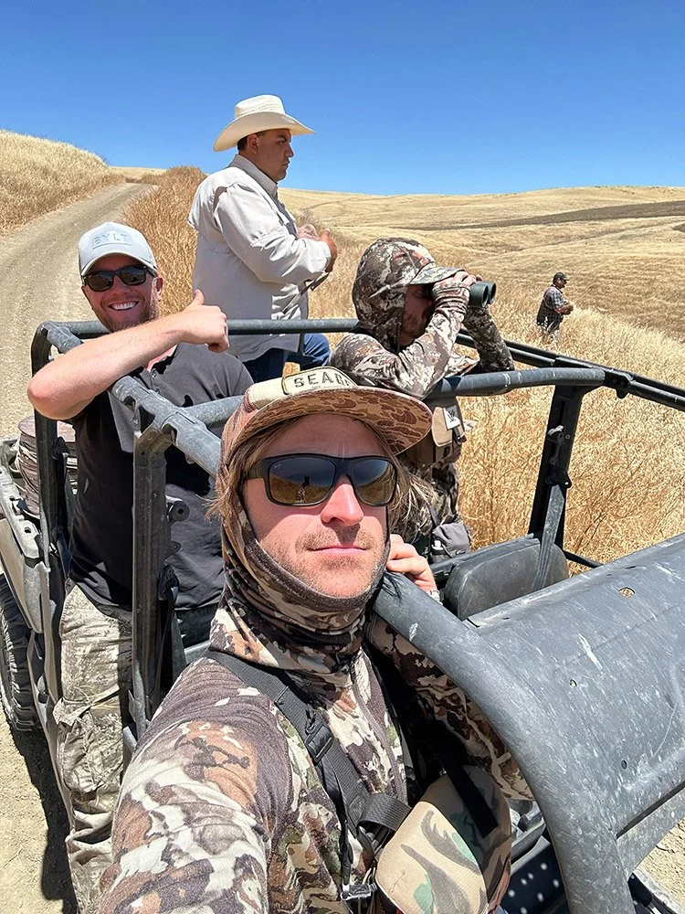 Four men on a rugged off-road vehicle in a dry, hilly field, with one man taking a selfie, two looking into binoculars, and one standing with arms crossed, wearing hats and camouflage gear.