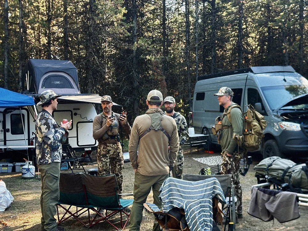 A group of five men in outdoor gear and camouflage, standing and talking near camping equipment and vehicles in a forest setting.