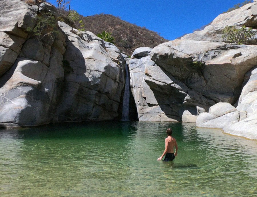 A man in black shorts standing in a clear green pool of water near a small waterfall surrounded by large gray rocks with a hill and blue sky in the background.