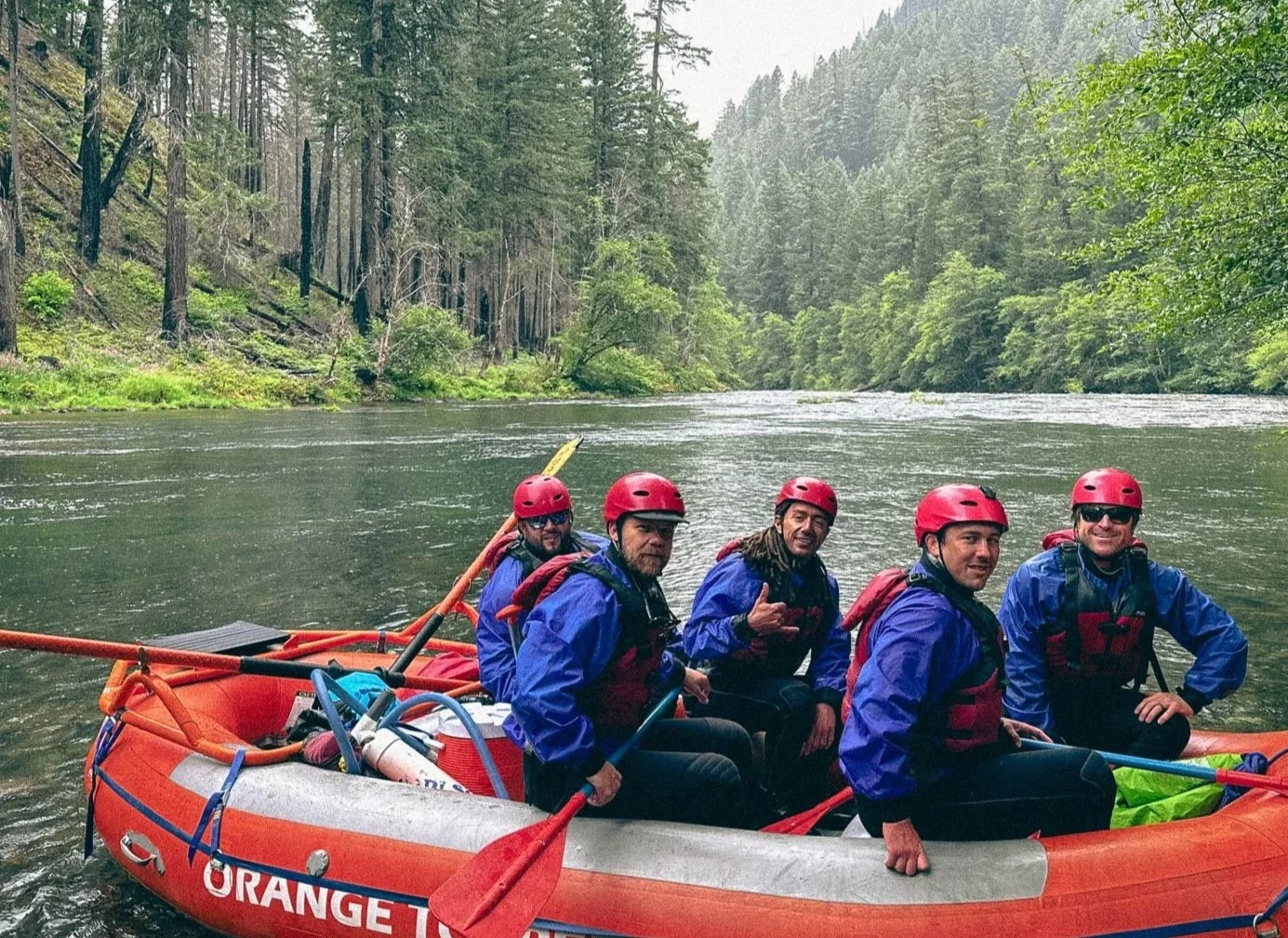 A group of five people wearing red helmets and blue life jackets sitting in an orange inflatable raft on a river surrounded by lush green trees.