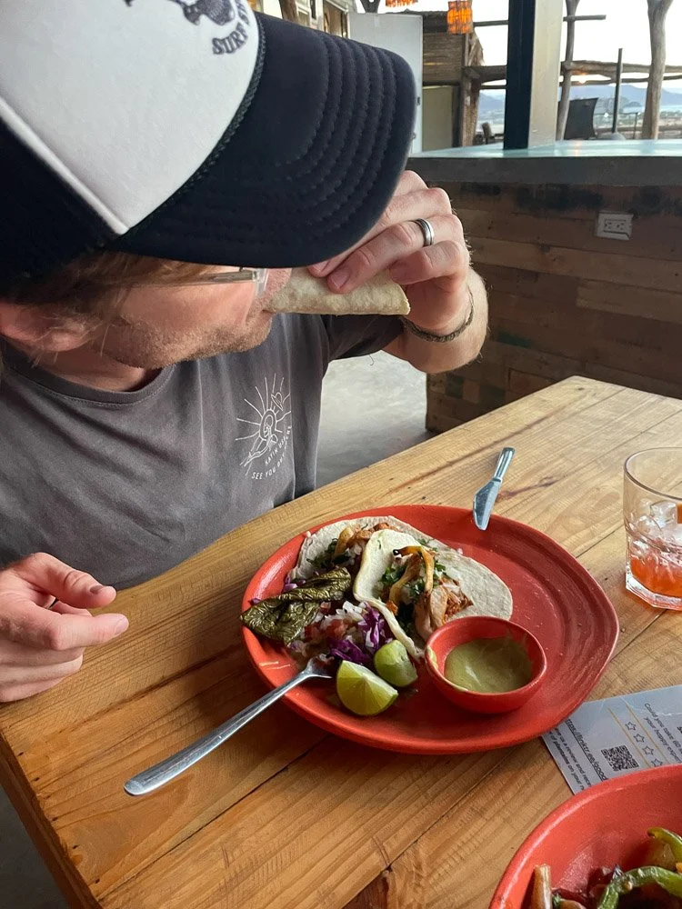 A man with a baseball cap eating a taco at a wooden table. There are additional tacos, lime wedges, and a glass of drink on the table.