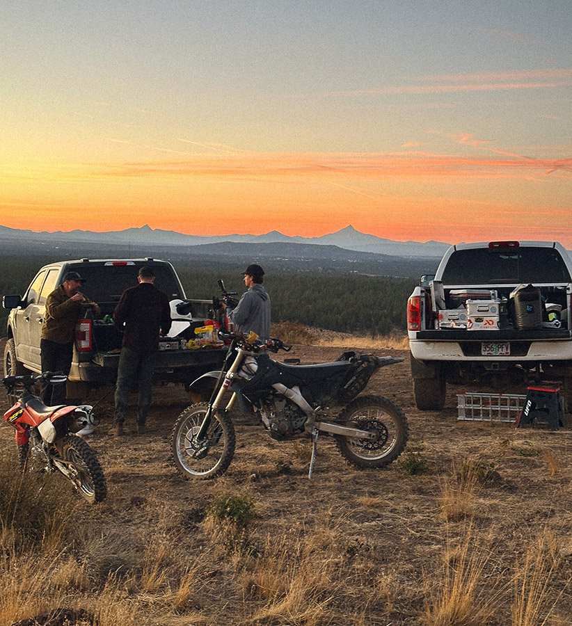 Three people are gathered around the open tailgate of a pickup truck on a dirt field during sunset. Two motorcycles are parked in front, and a second pickup truck is nearby with equipment in its bed. The background shows a scenic view of mountains an
