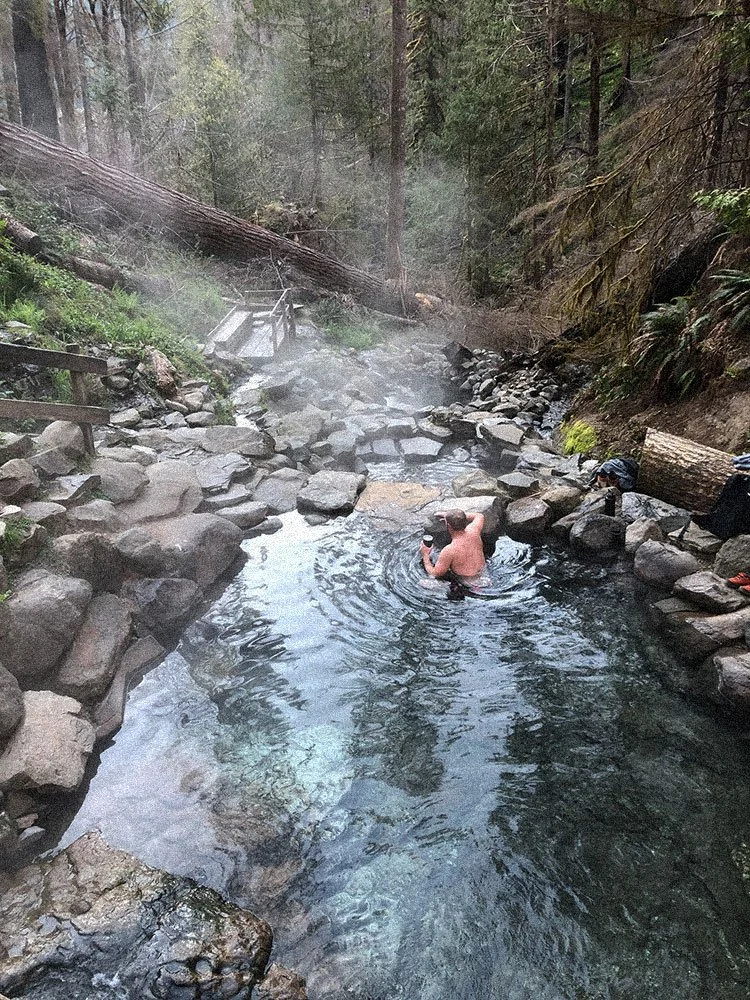 A man swimming in a natural hot spring surrounded by rocks and forest in a remote area.