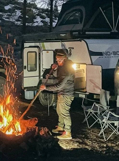 Person roasting marshmallows over a campfire next to a camper trailer and foldable chairs during dusk.