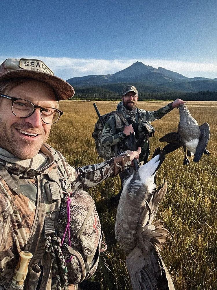 Two men in camouflage clothes and hats standing in a field with mountains in the background, holding hunting gear and three ducks, one of which they are displaying.