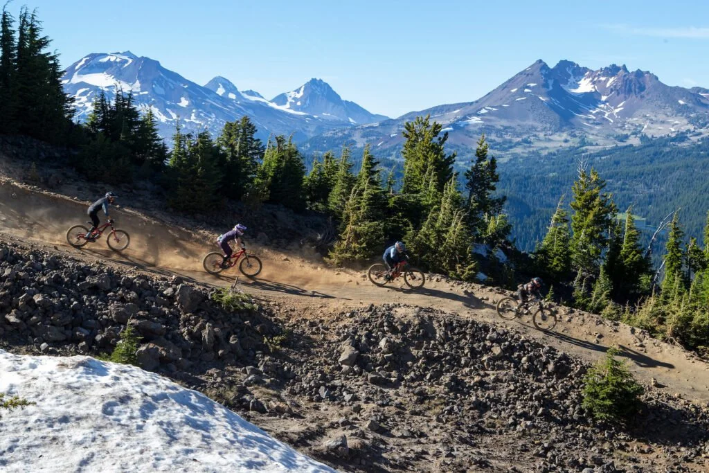 Four mountain bikers riding on a dirt trail through a mountainous landscape with snow-capped peaks and green pine trees.