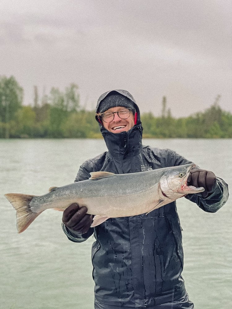 Person in rain gear holding a large fish on a cloudy day by a body of water.